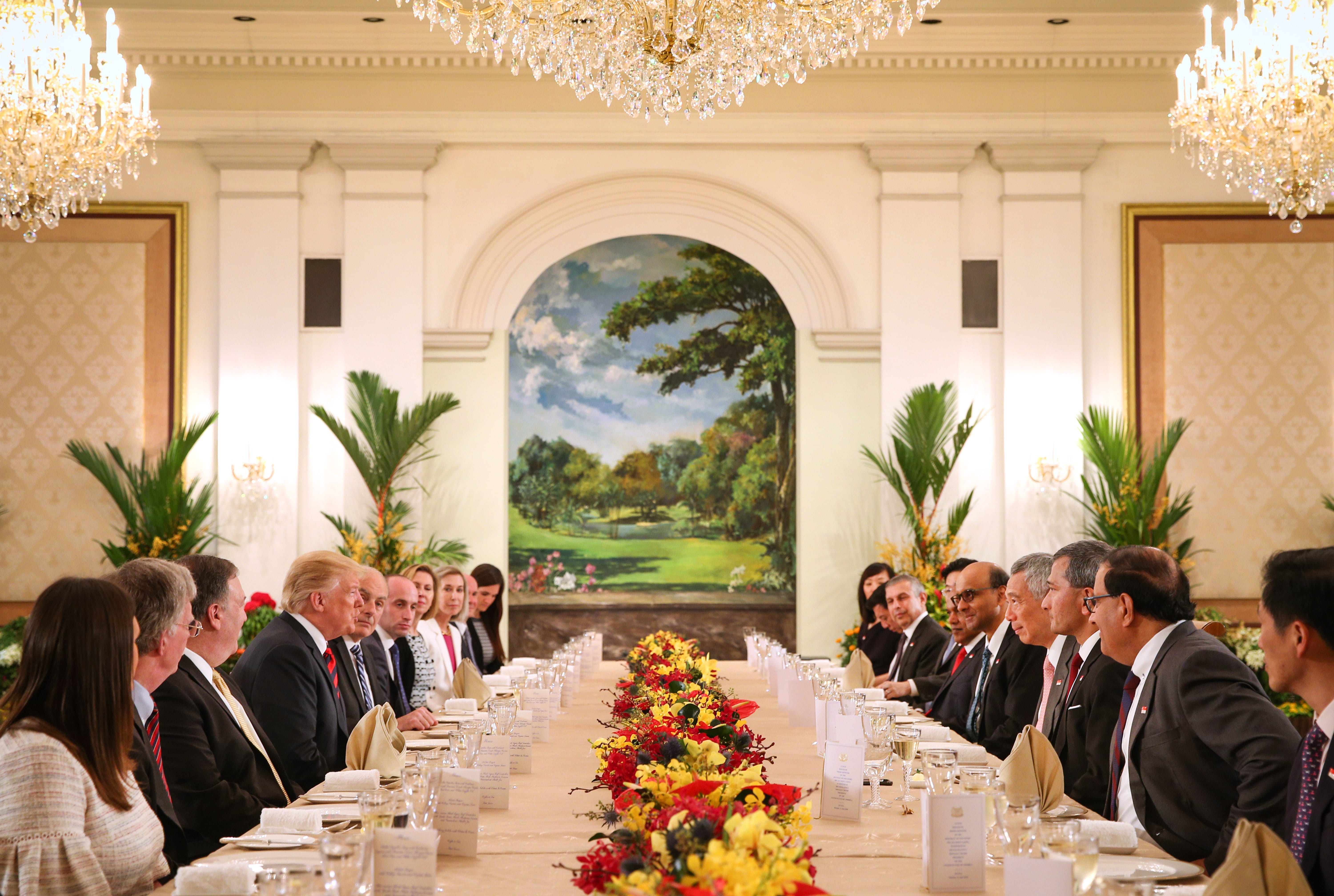 Donald Trump & others at a formal dinner table in a bright room with chandeliers & a mural.