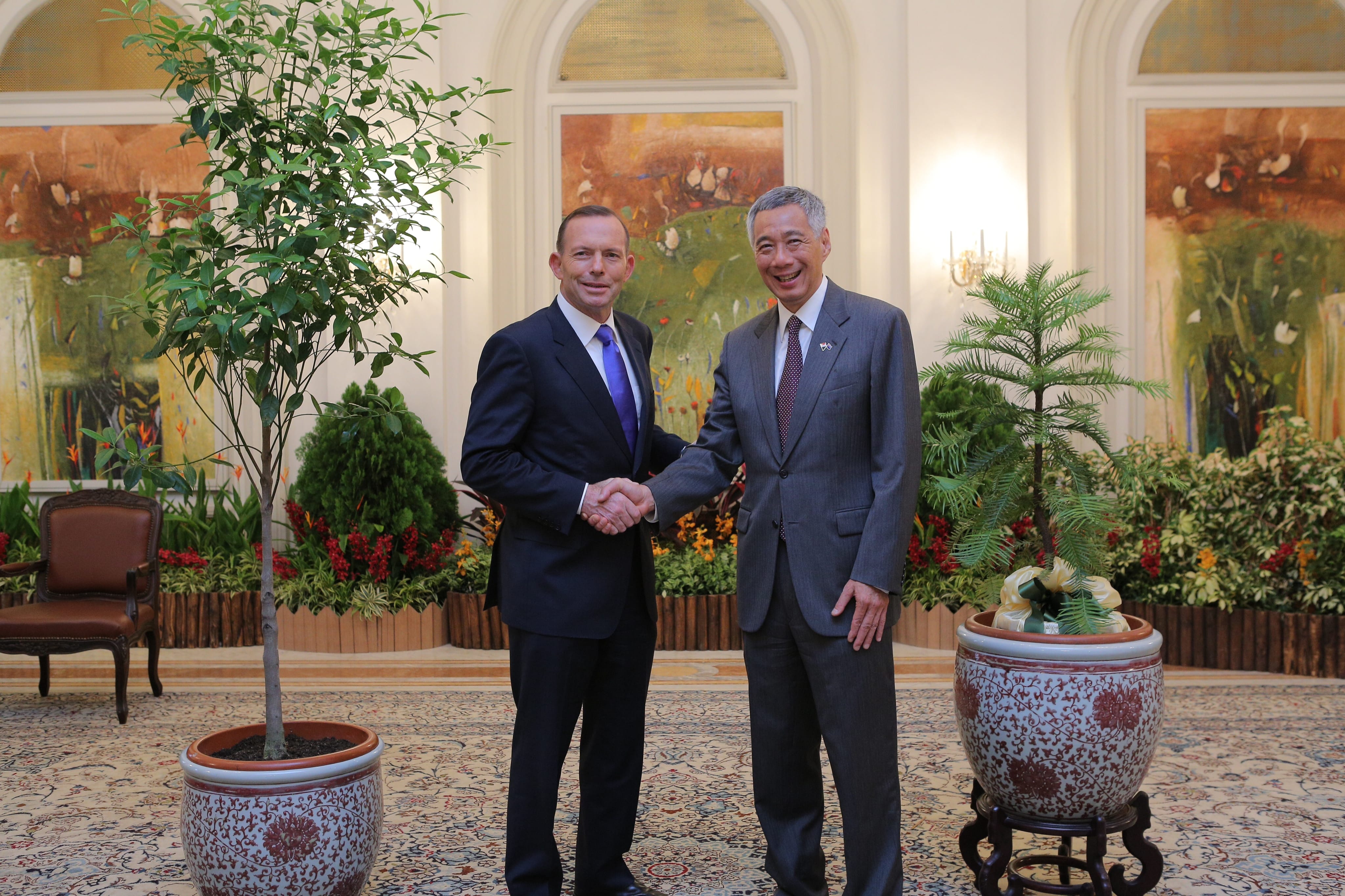 Two men in suits, Tony Abbot and Lee Hsien Loong, shake hands in a room with potted plants and paintings.