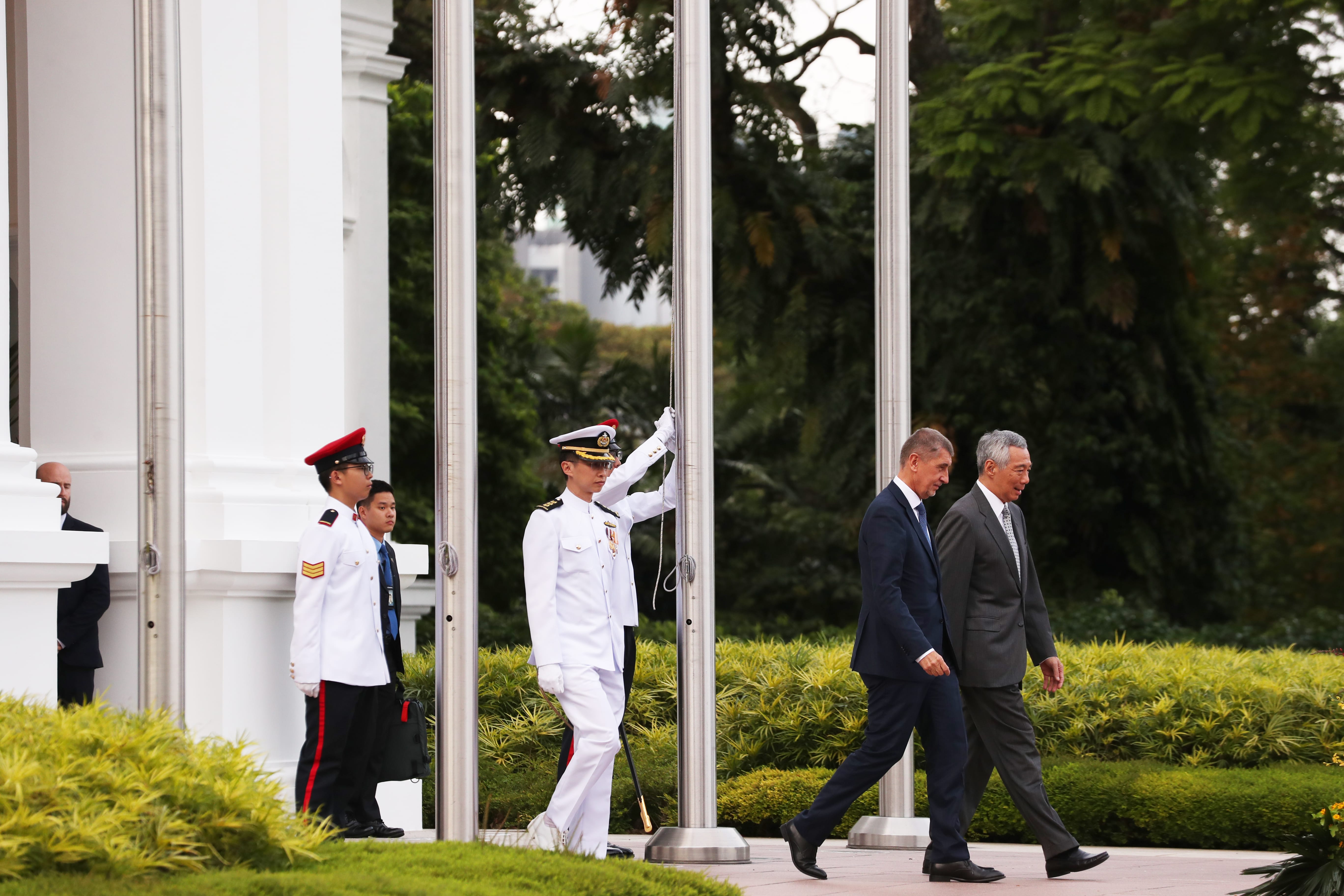 Two men in suits walk past uniformed officials near flag poles on a paved path.