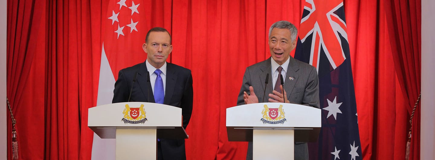 Two men at podiums before flags of Singapore and Australia against a red curtain.