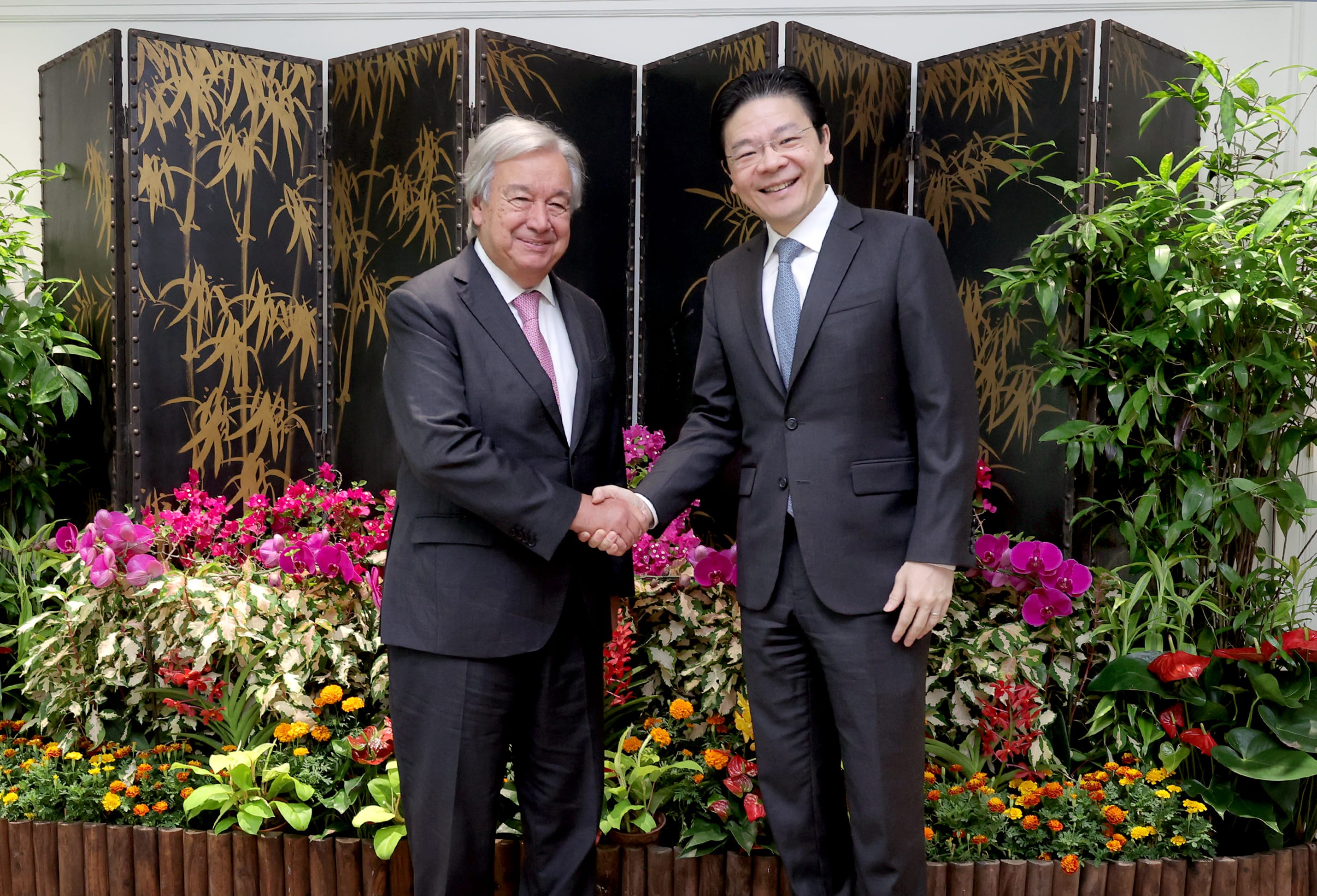 António Guterres and a man shake hands in front of a floral display and gold bamboo screen.