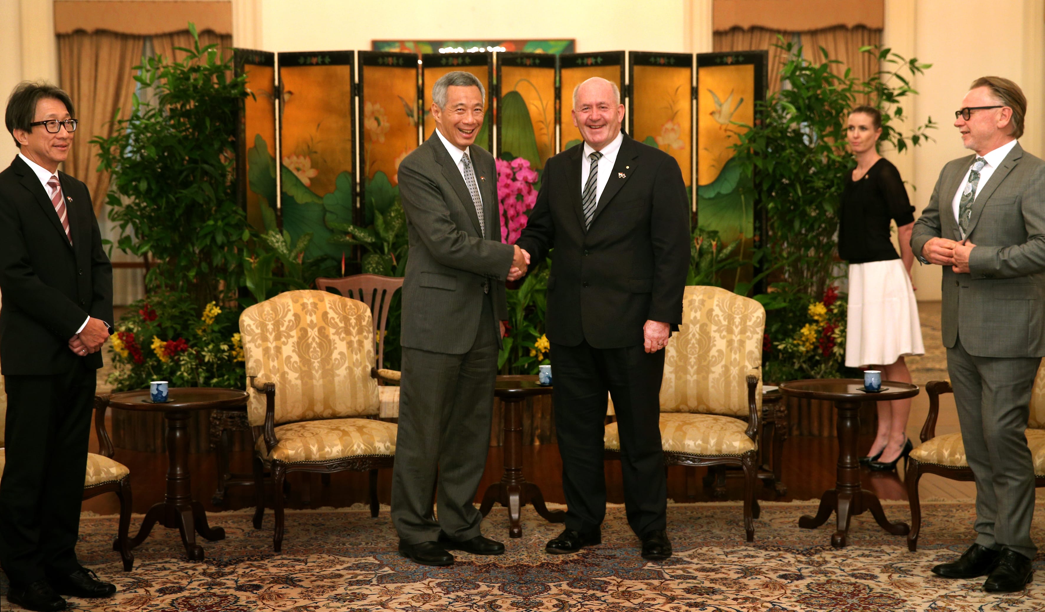 Two men in suits shake hands in a room with decorative screens and patterned carpet.
