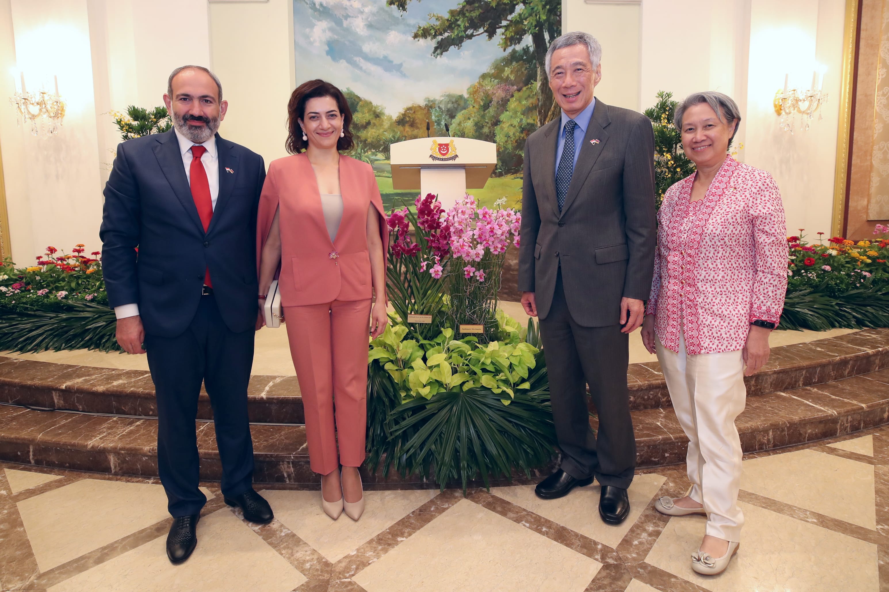 Four people stand in formal attire before a podium with an emblem and floral arrangements.