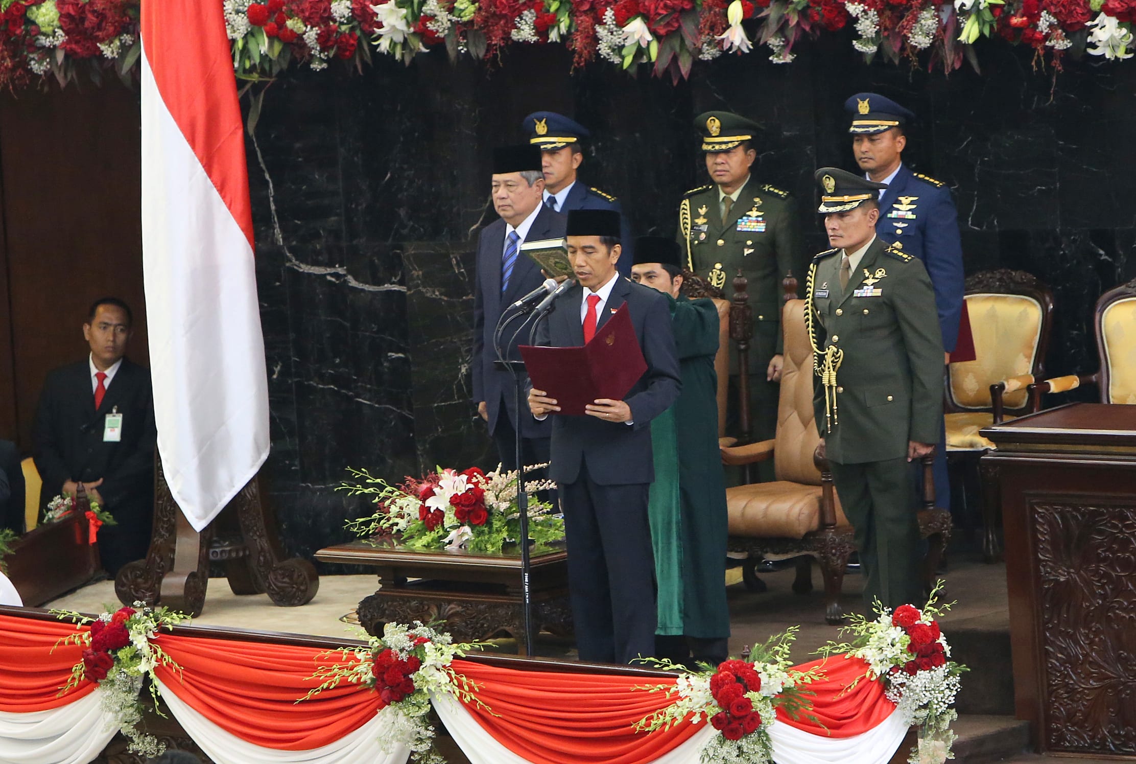 Jokowi reciting oath with military officials, red/white flag, and flower decorations.