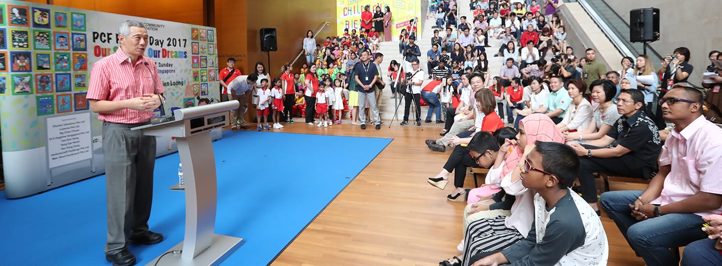 Lee Hsien Loong speaks at a podium before a seated crowd with children standing at the side.