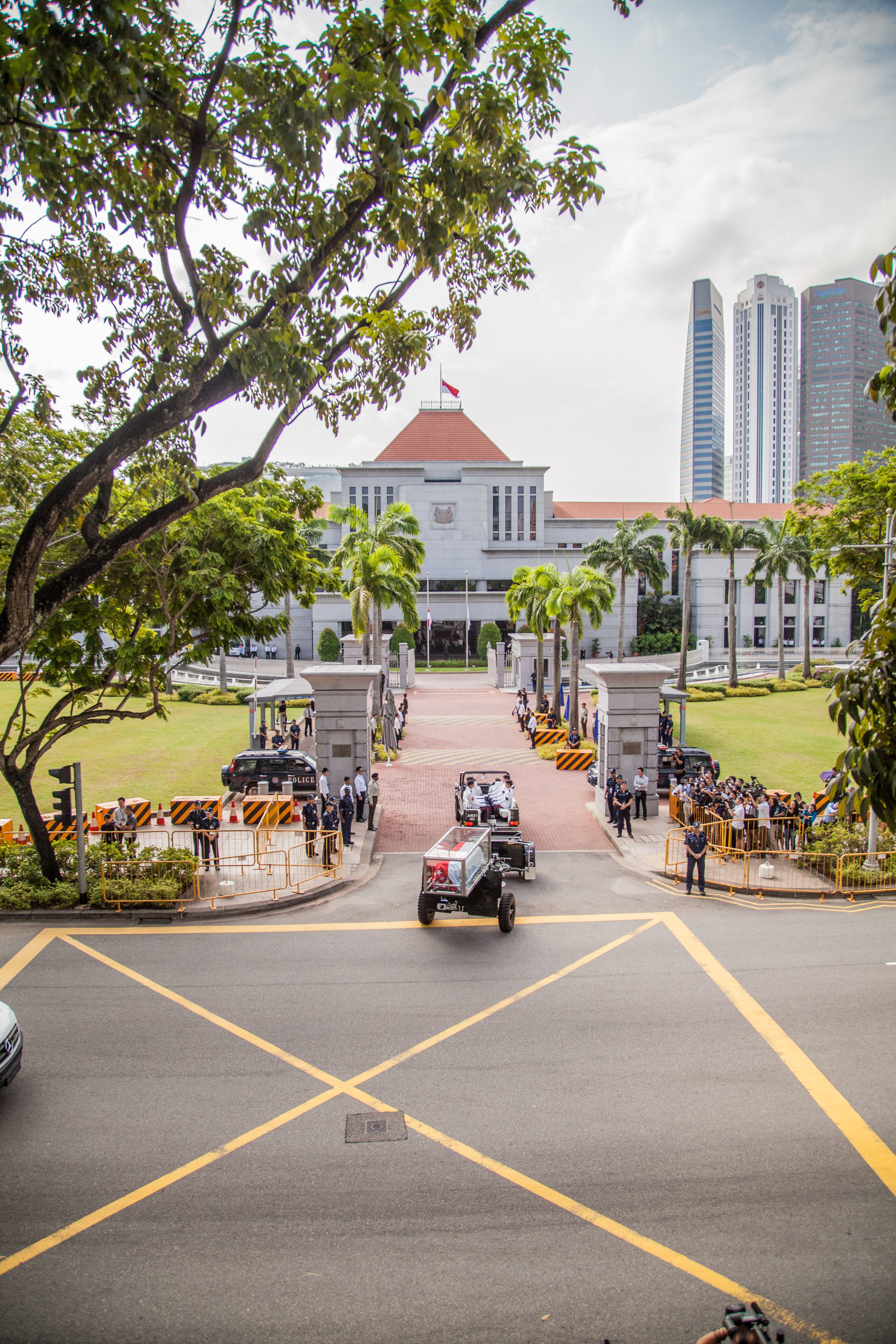 Ceremonial vehicles with coffin approaching building lined with people and palm trees. Tall buildings in the background.