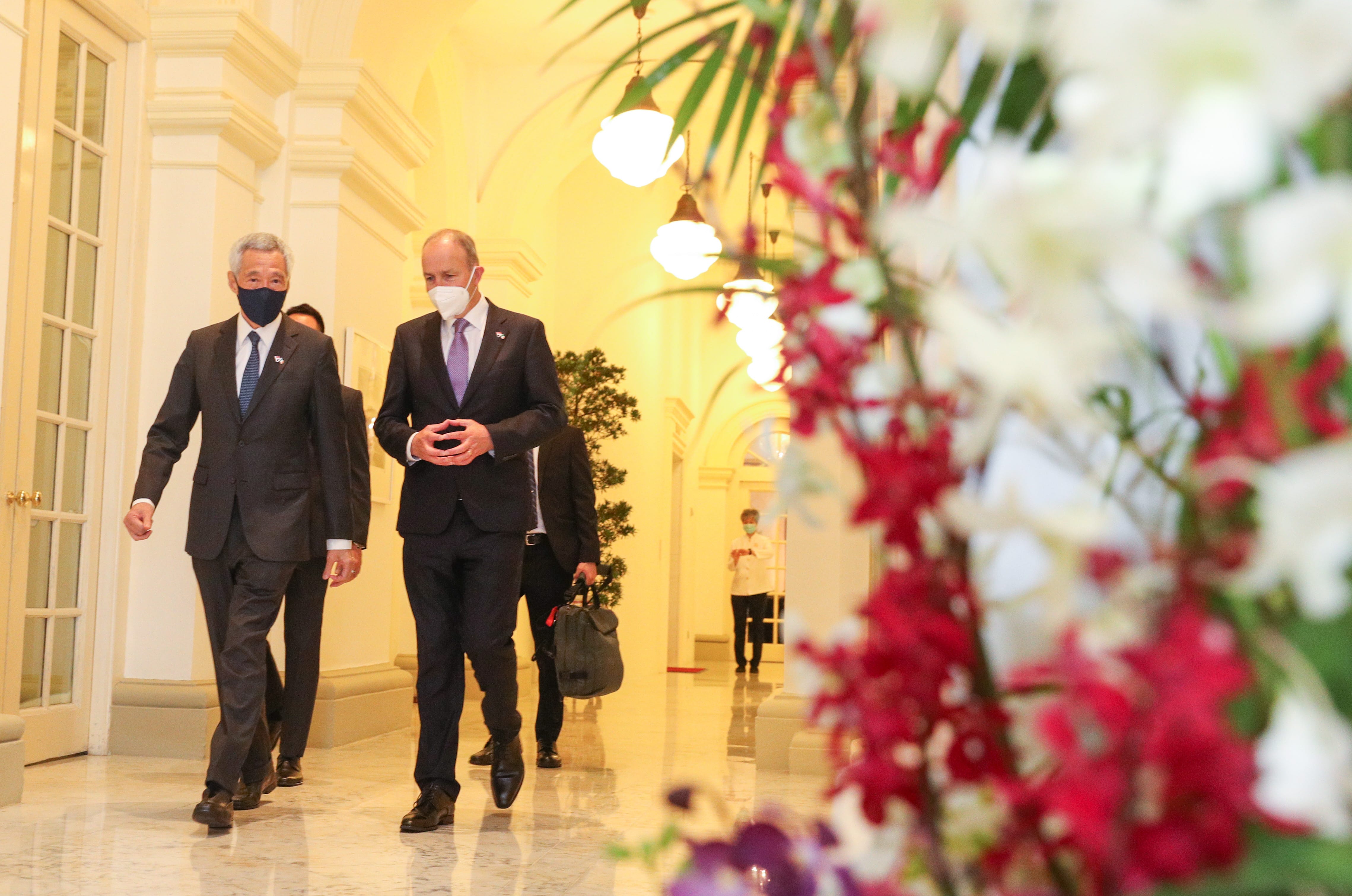 Two men in suits and masks walk down a hallway decorated with flowers and ornate lighting.