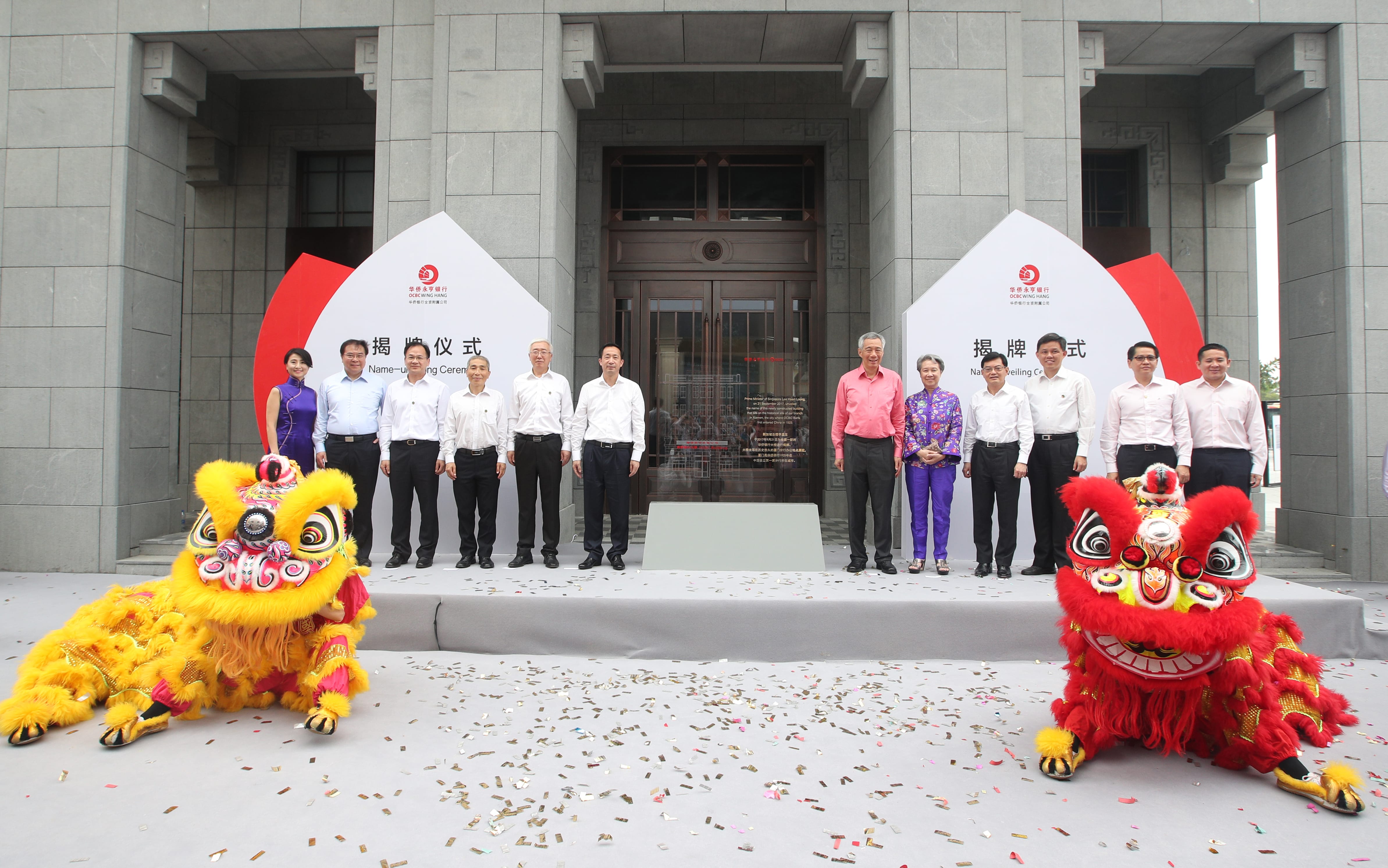 Group poses before OCBC Wing Hang building; confetti and two lion dance costumes present.