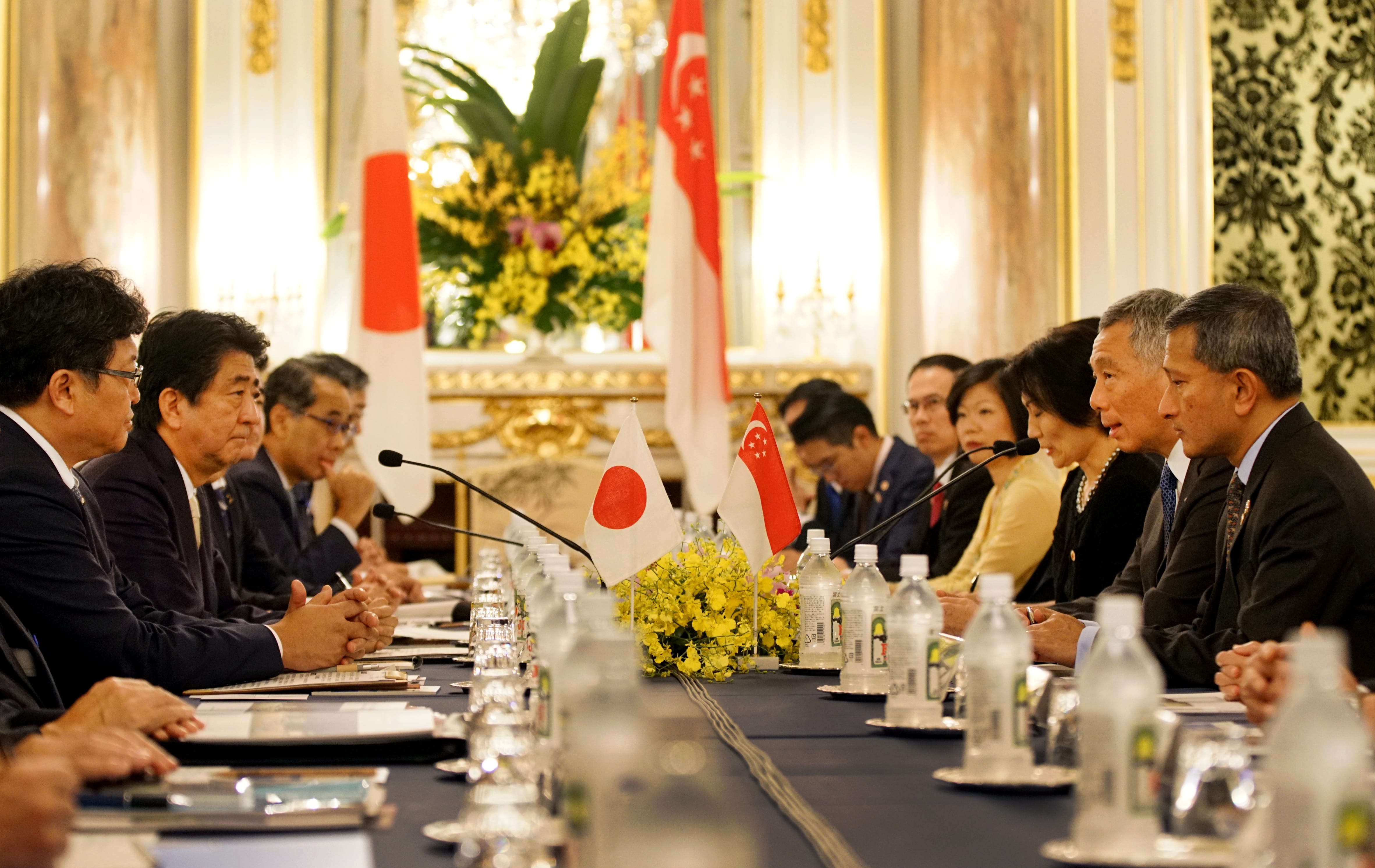 People at long table with Japan and Singapore flags, water bottles, and microphones.