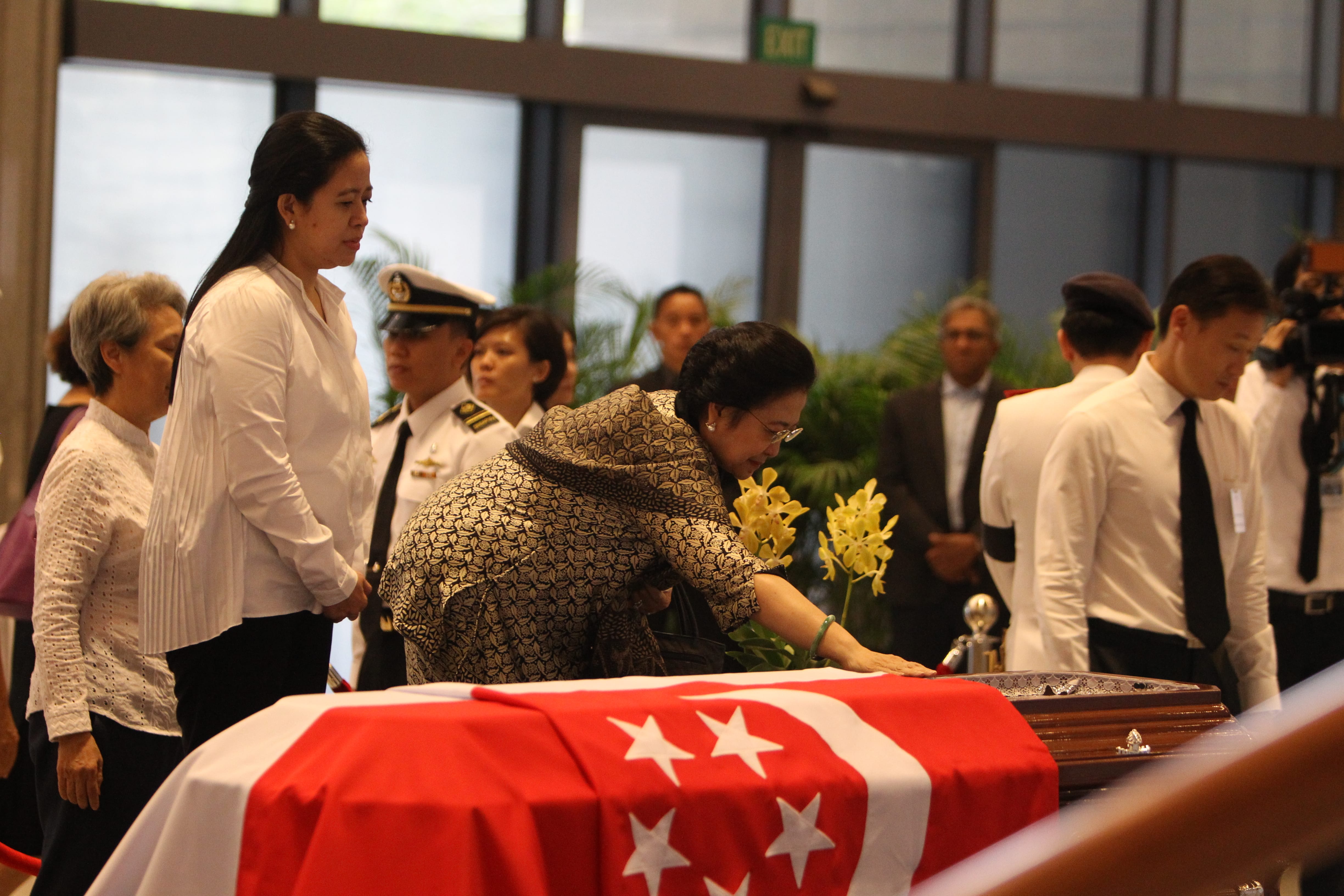 Woman touches a casket draped with the Singapore flag, with yellow flowers and onlookers.