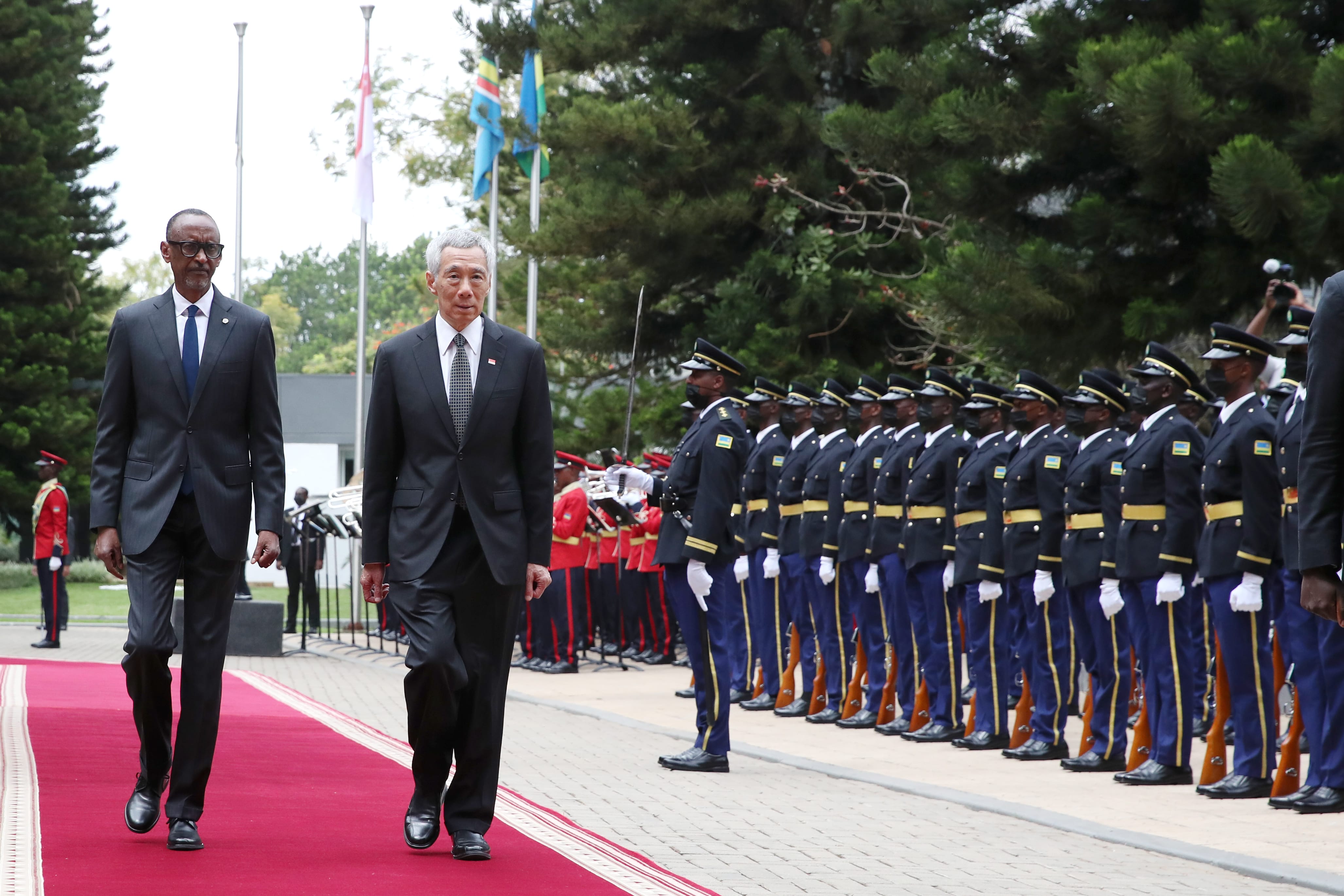 Two men in suits walk on a red carpet past a military honor guard with rifles.