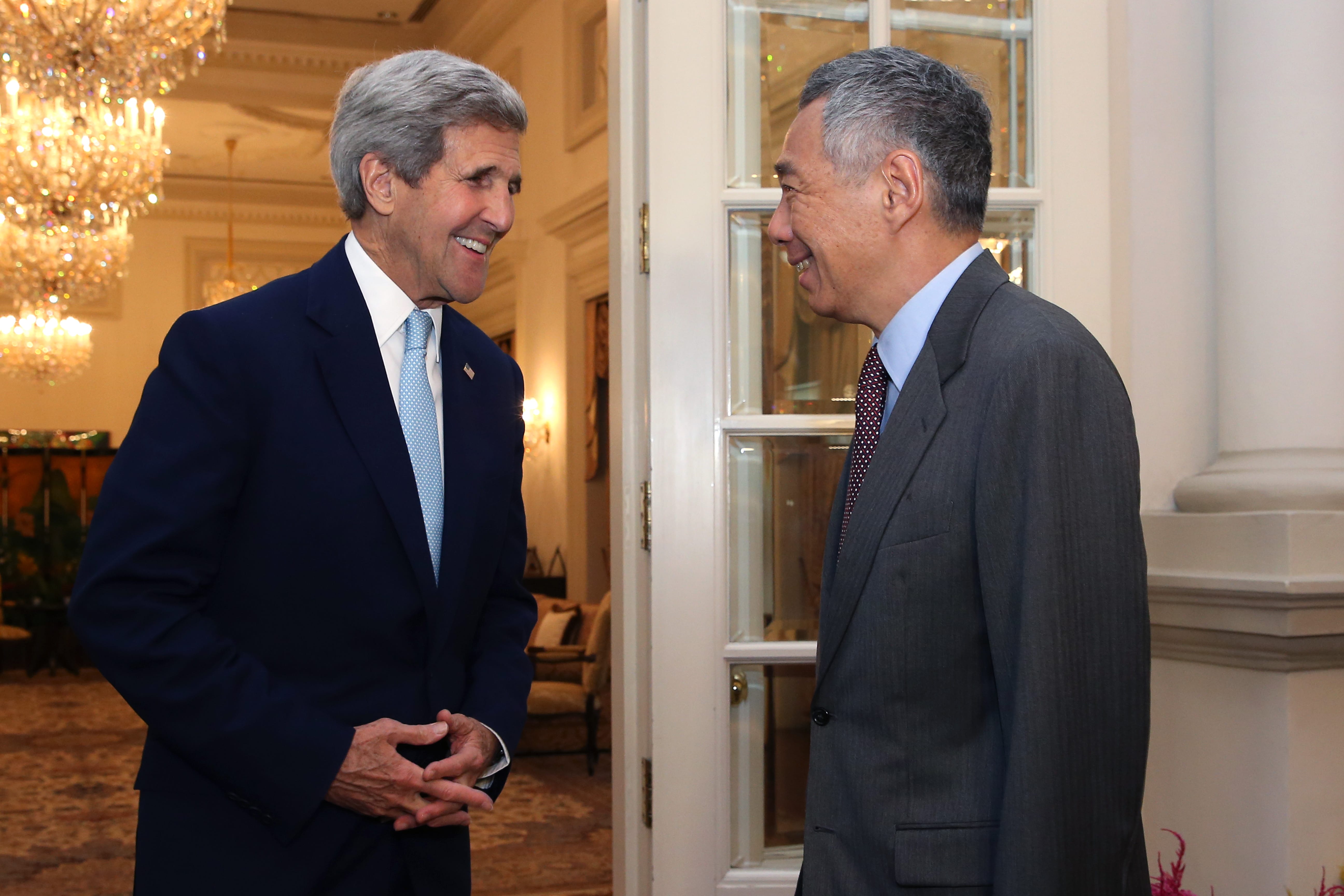 John Kerry and Lee Hsien Loong face each other, smiling in suits, standing in a doorway.