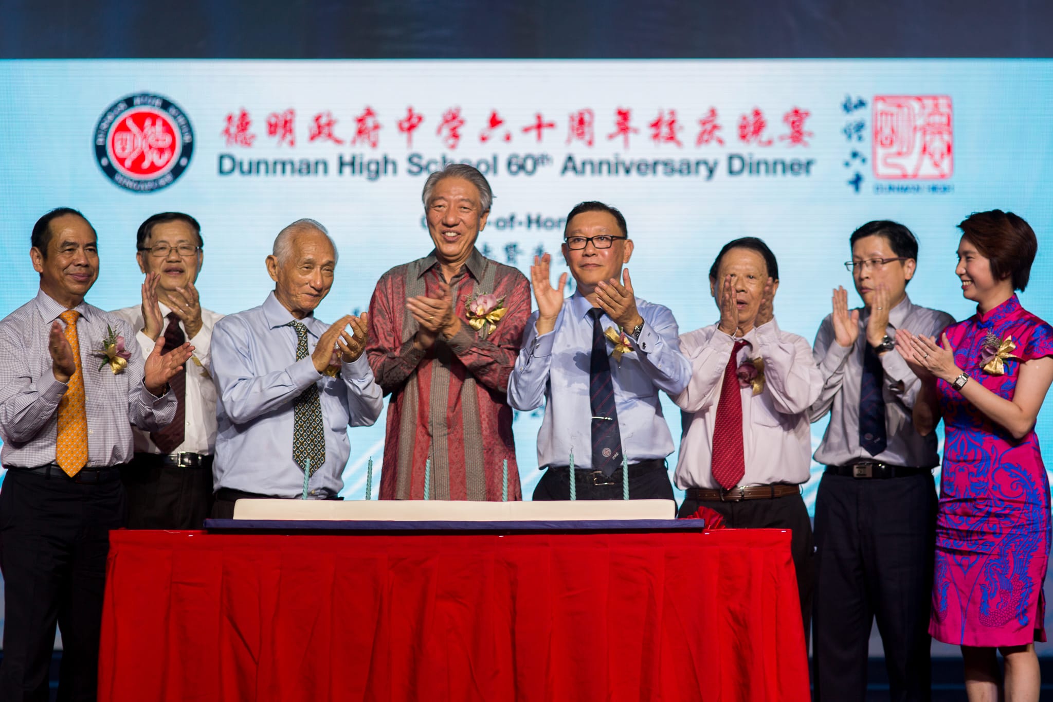 Group of people applauding behind a cake on a red table. Dunman High School 60th Anniversary Dinner backdrop.