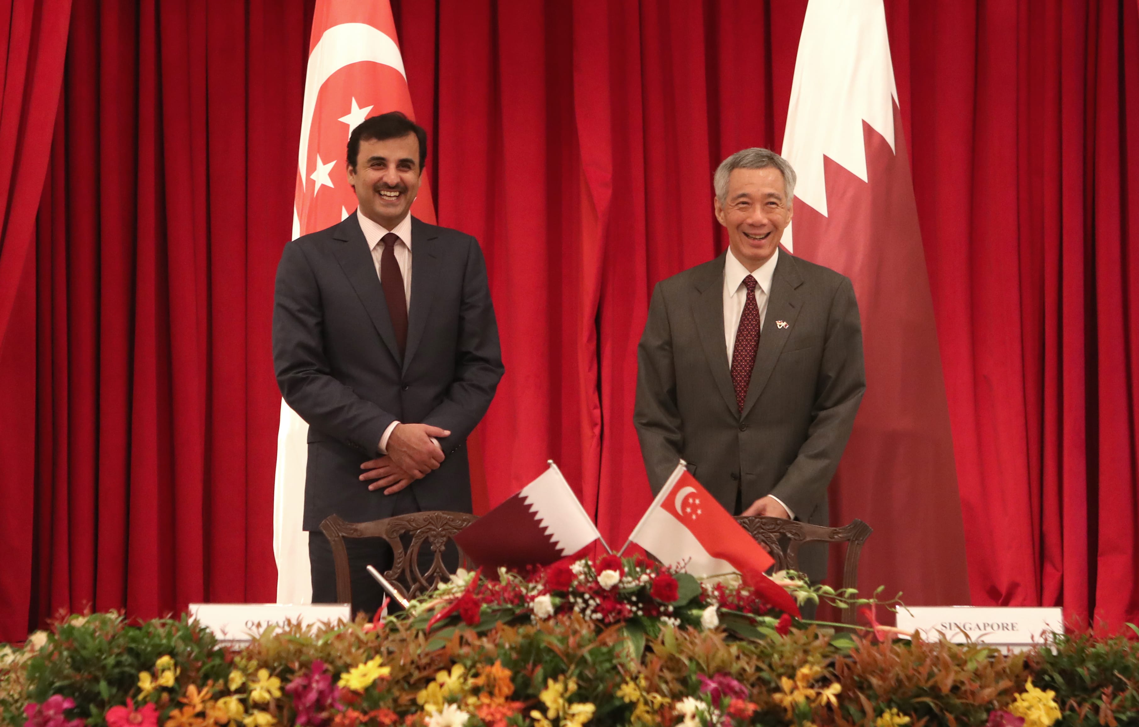 Two men in suits stand before Qatar and Singapore flags, above a floral arrangement.
