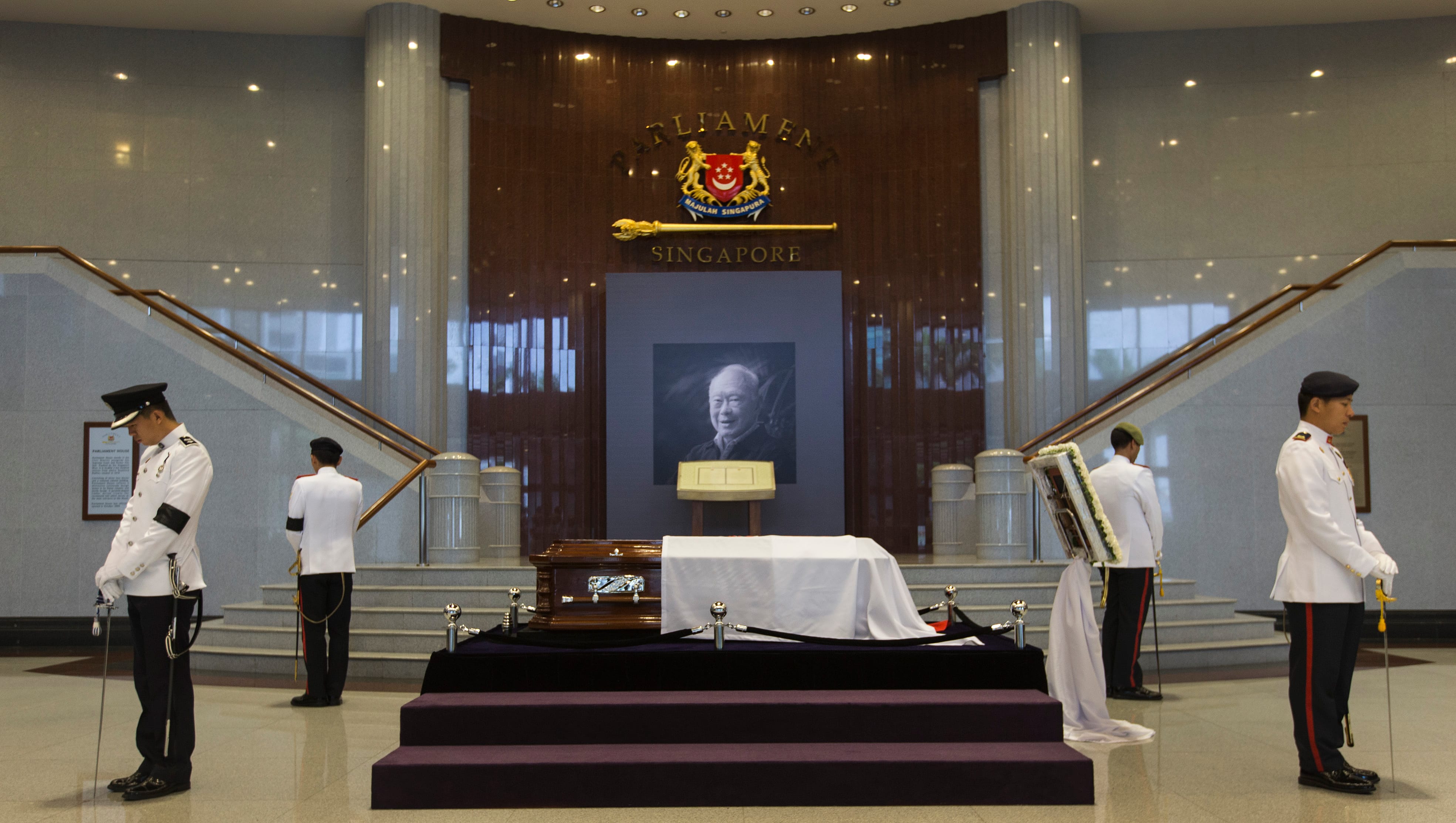 Ceremonial guard in dress uniform flanking a coffin, under a portrait. Singapore Parliament logo visible.
