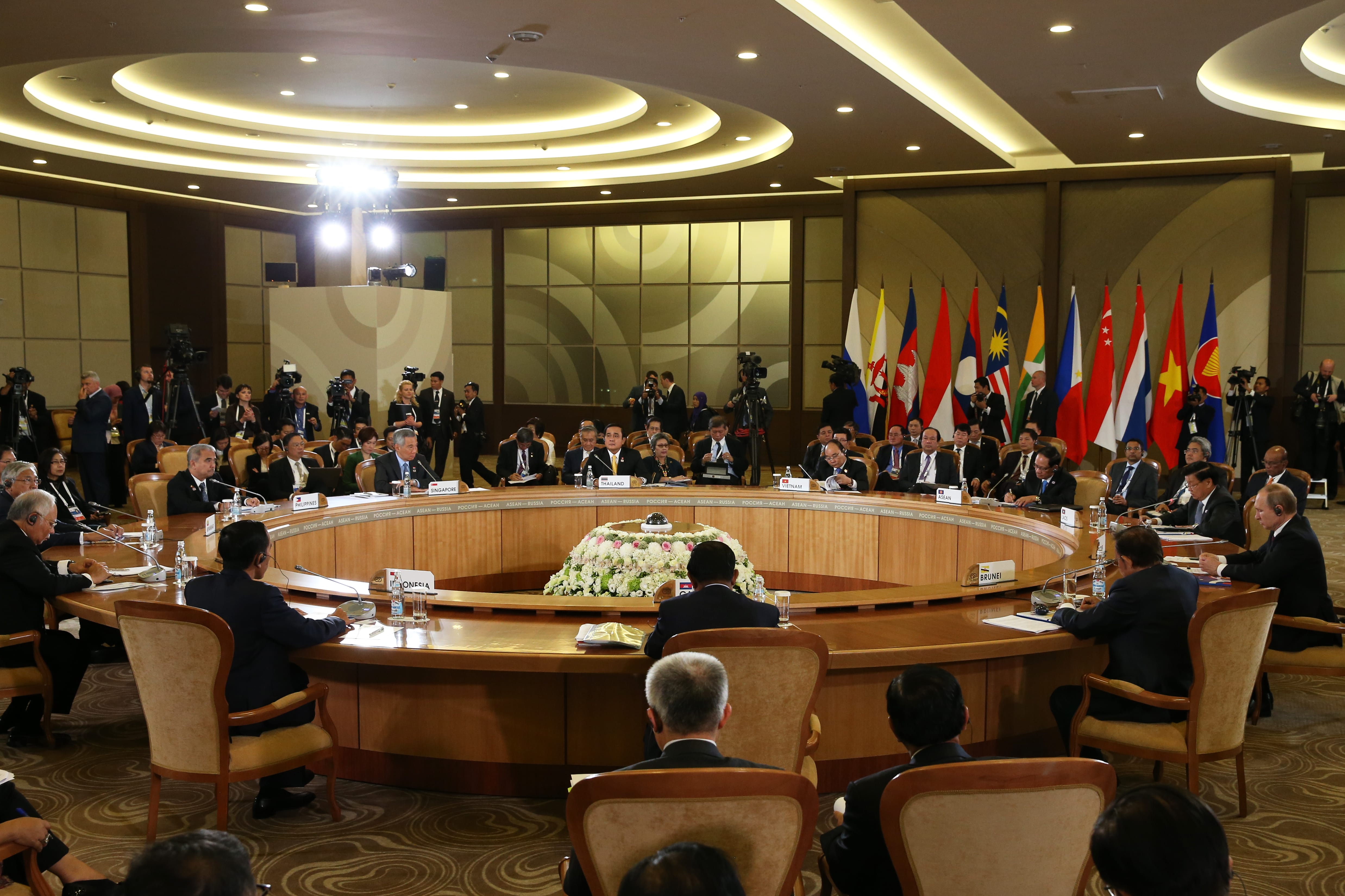 Delegates seated at round table beneath flags, press behind.