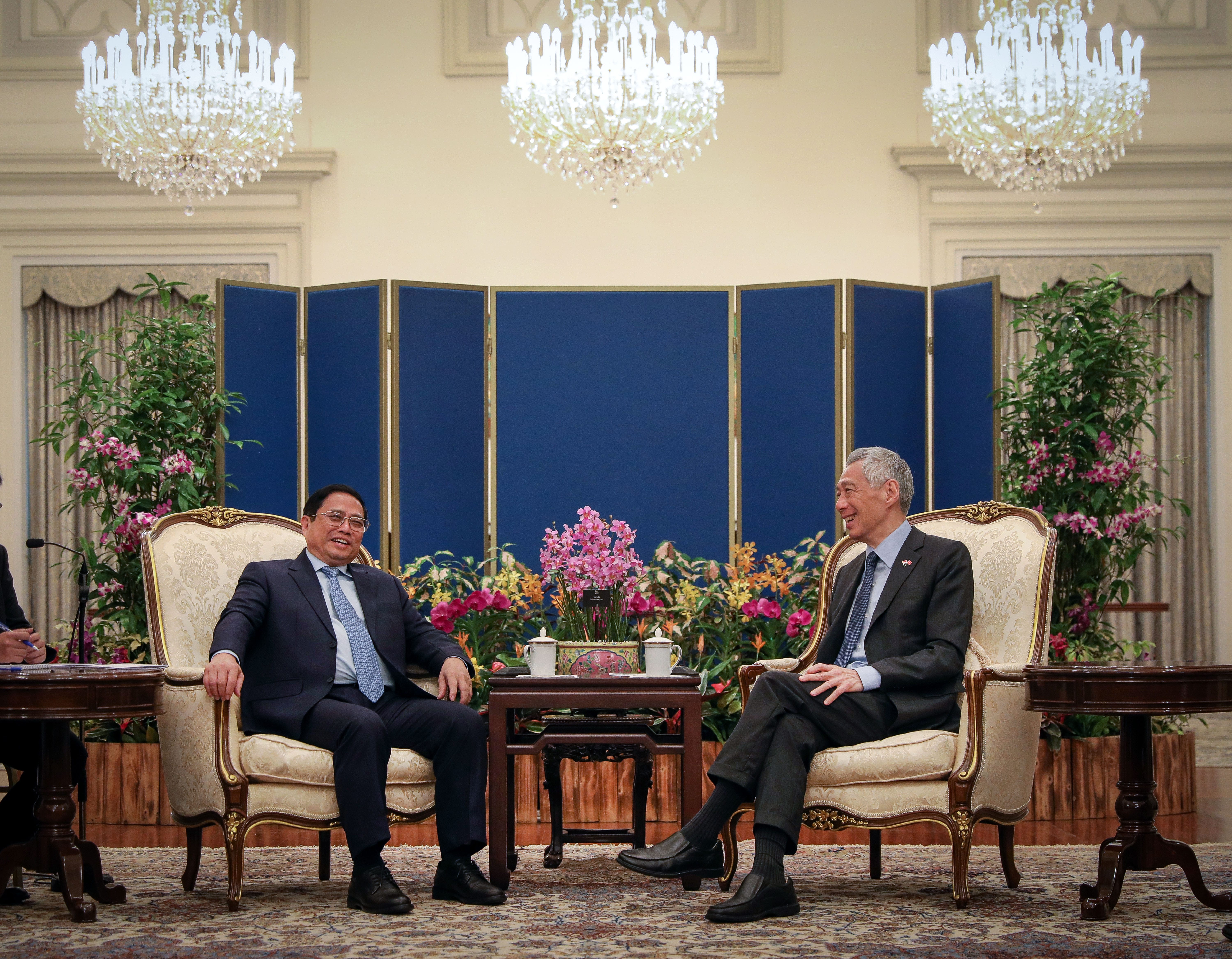 Two men in suits seated indoors, with crystal chandeliers and flower arrangements.