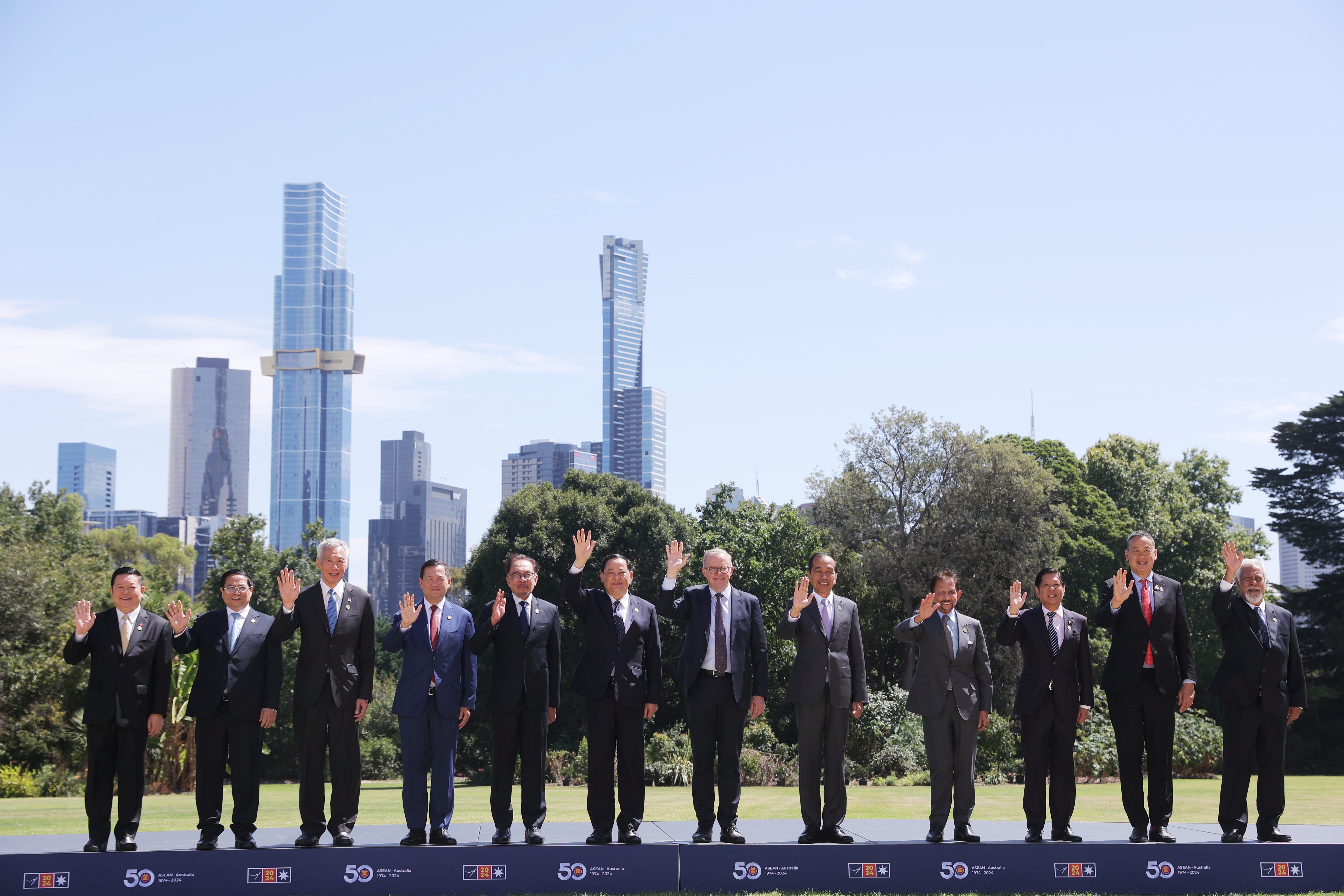 Eleven people in suits wave in front of the Melbourne skyline. ASEAN-Australia 1974-2024 logo at base.