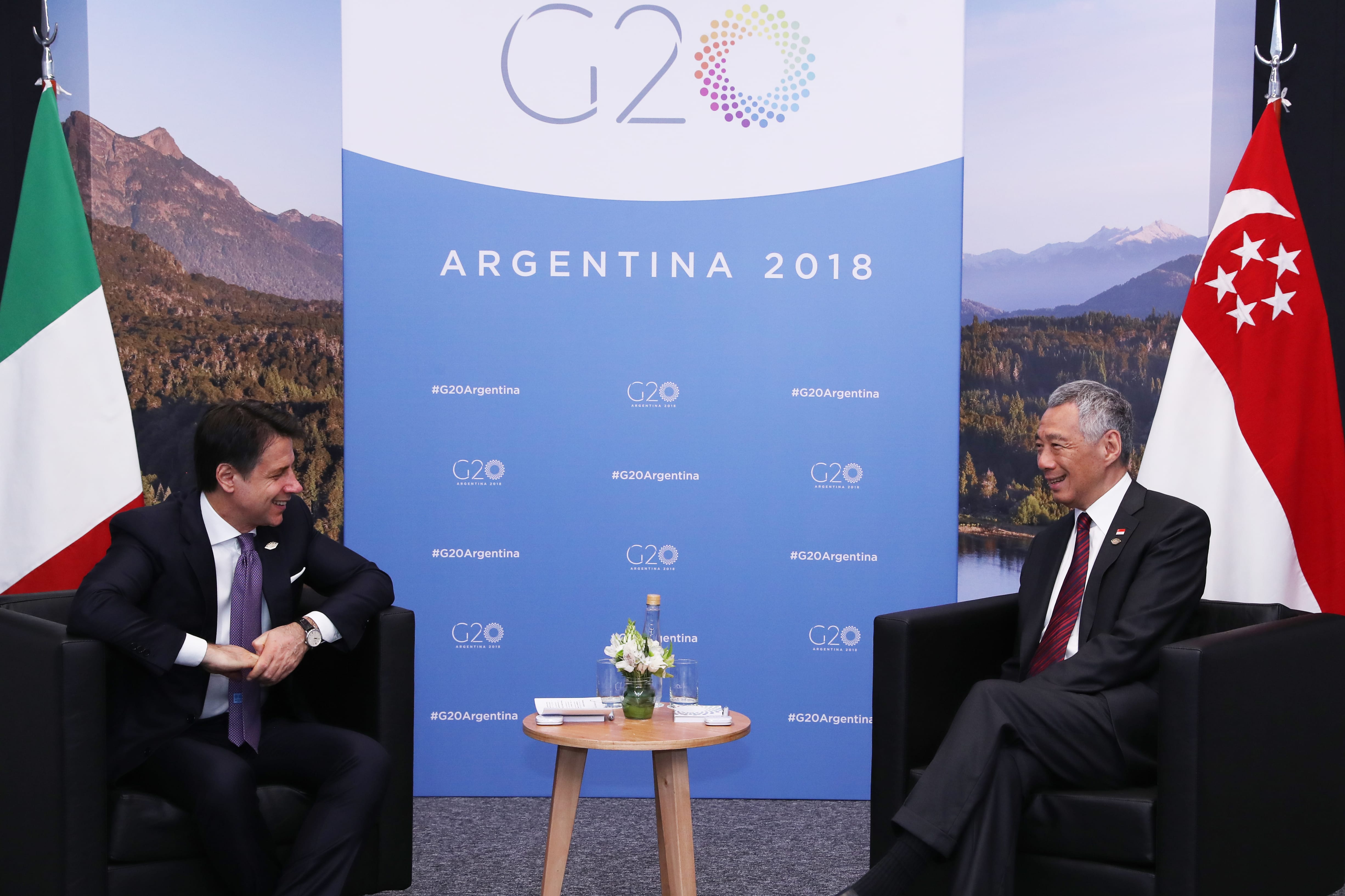 Leaders sit before a G20 Argentina 2018 backdrop, next to Italian and Singaporean flags.