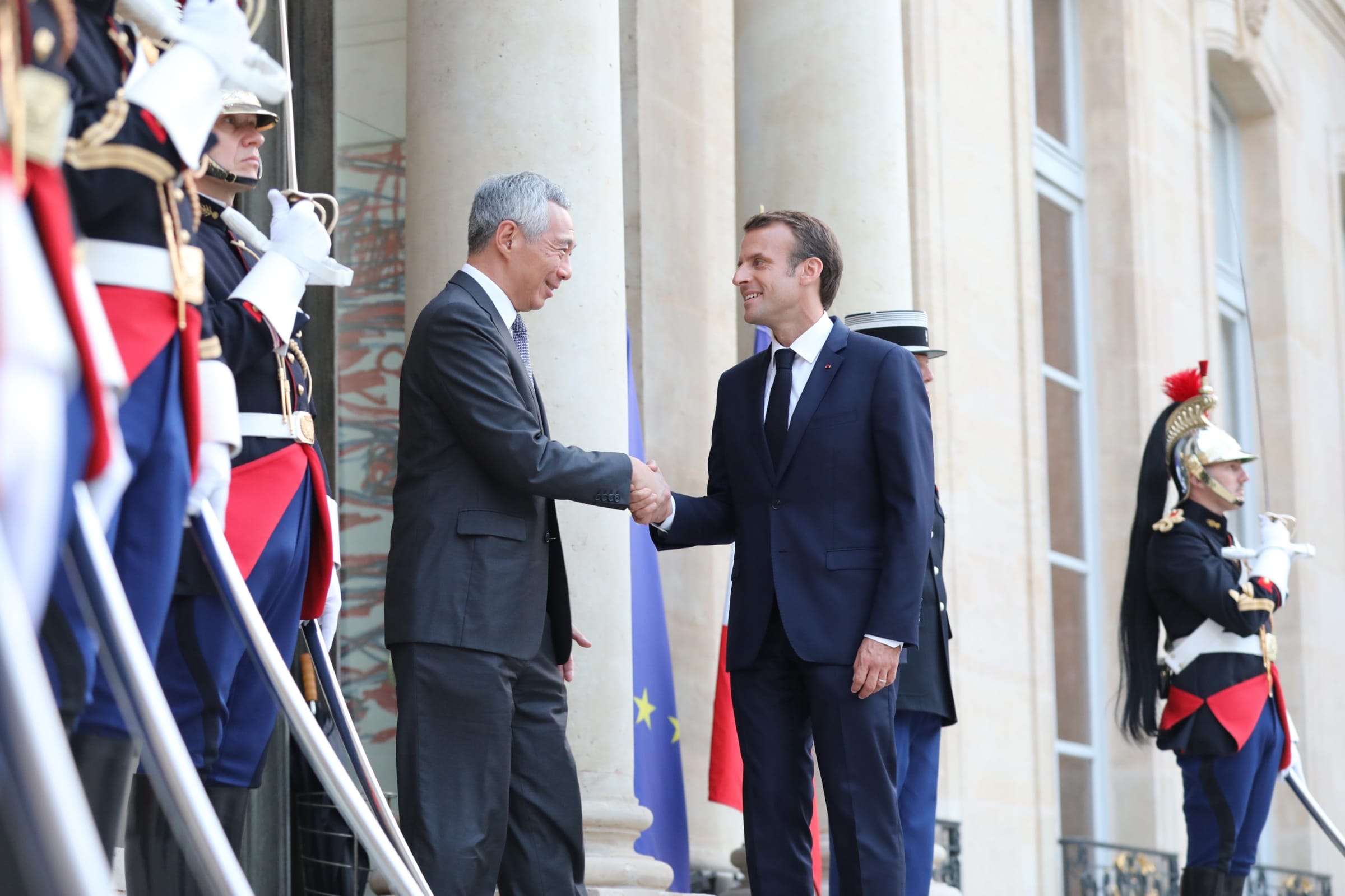 Lee Hsien Loong and Emmanuel Macron shake hands before guards in formal attire and the French & EU flags.