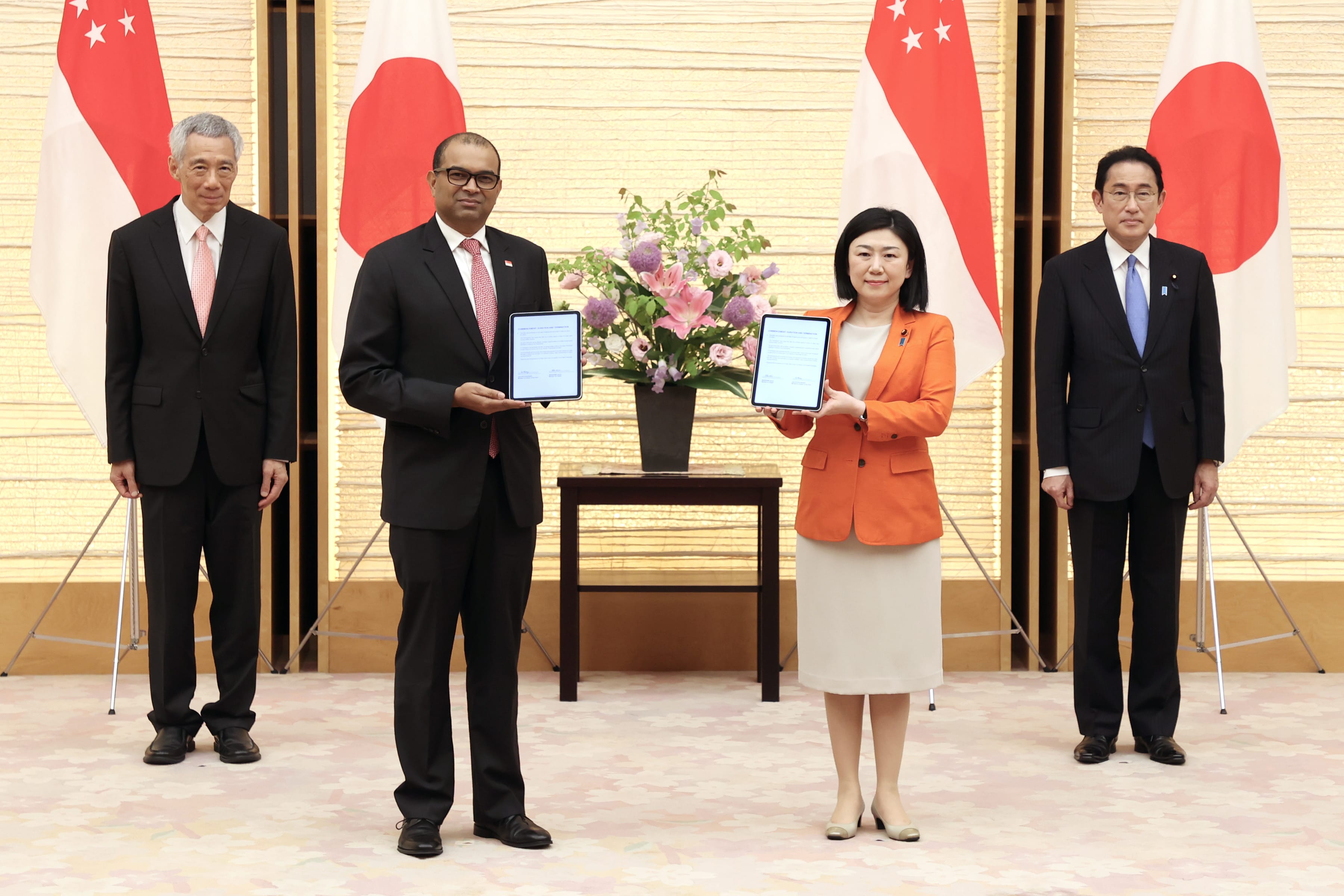 Four people hold tablets, standing before Singapore and Japan flags with floral arrangement.
