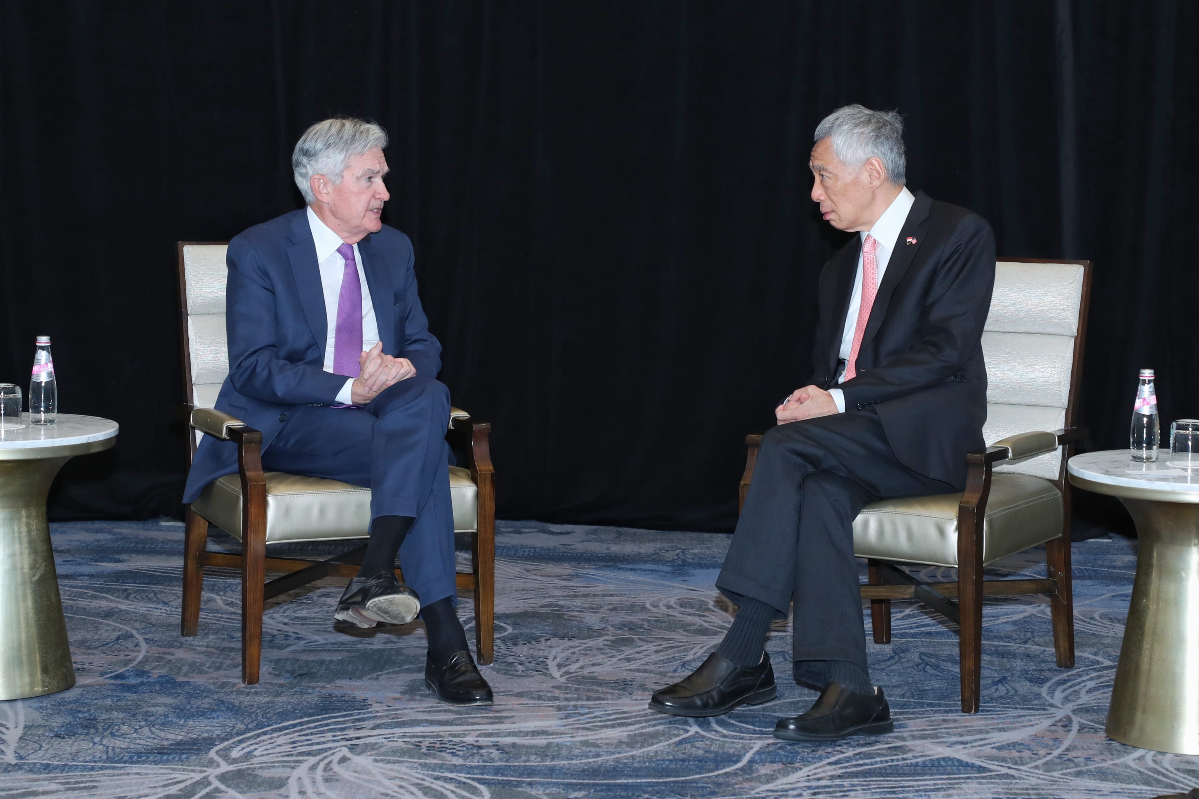 Two men in suits seated, facing each other in upholstered chairs against a dark backdrop. Water bottles nearby.