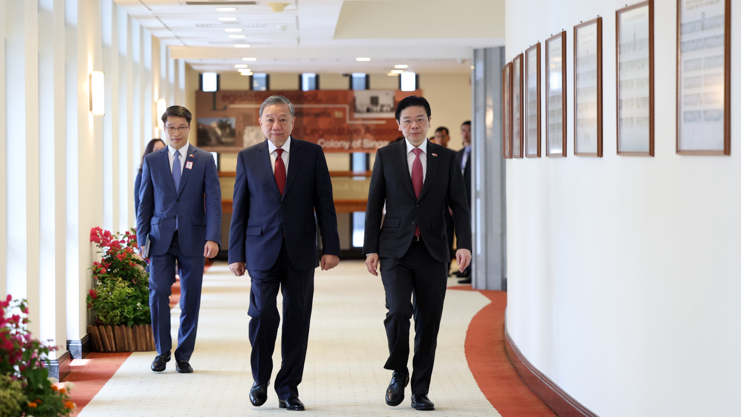 Three men in suits walk down a corridor with framed documents on the wall.