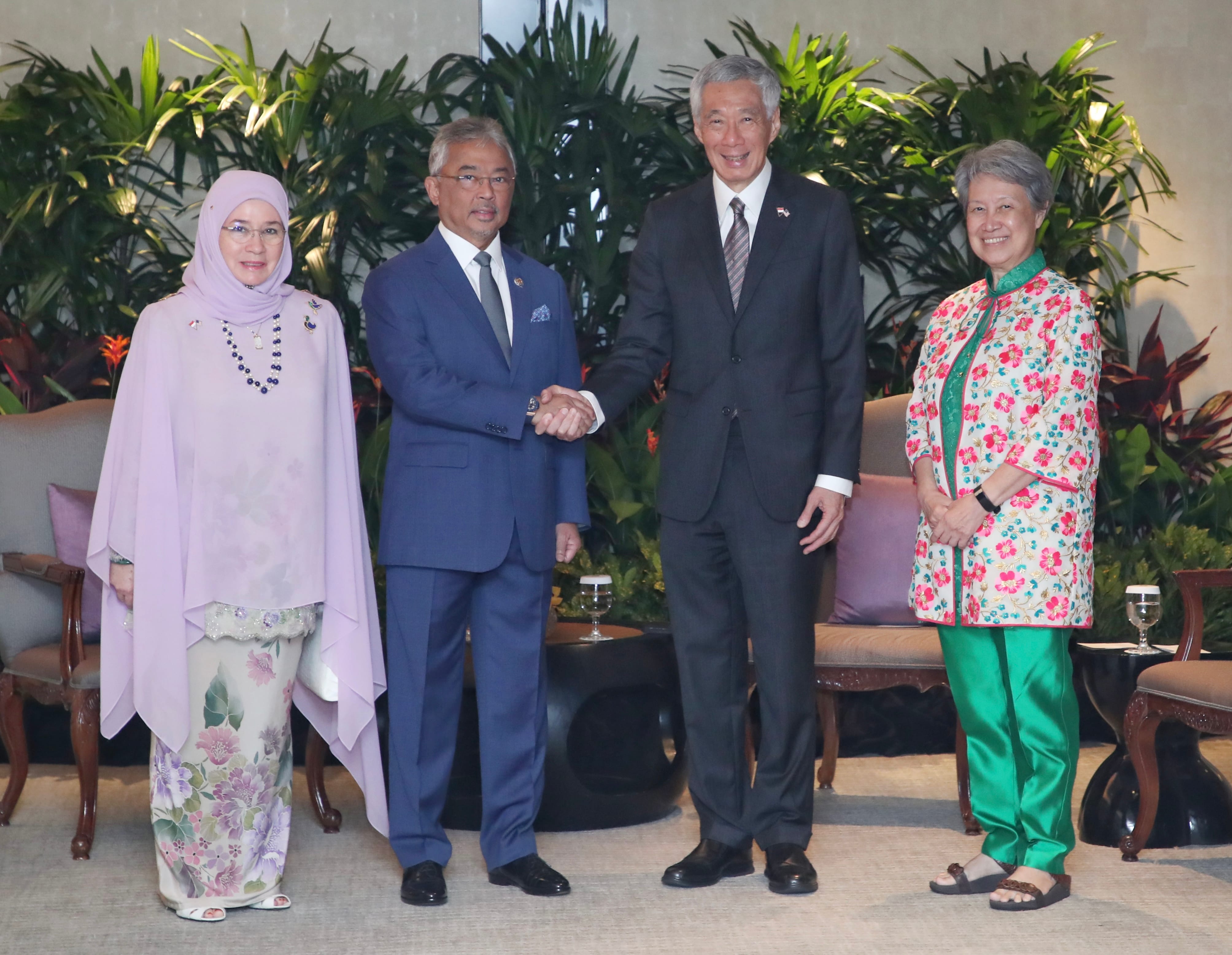Four people in formal wear shake hands in front of greenery. Lee Hsien Loong is in a suit.