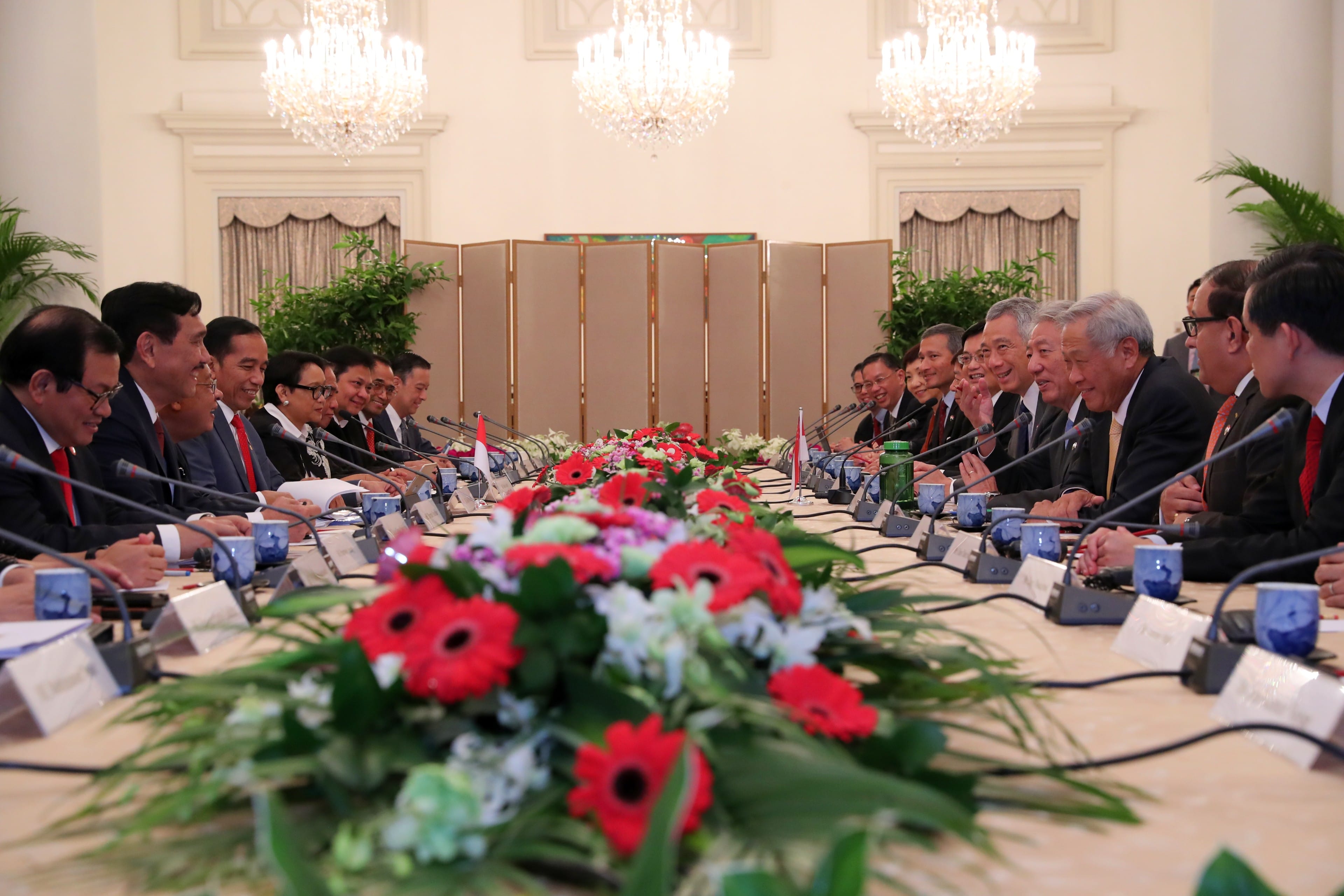 Formal meeting with rows of people behind microphones at a long table with floral centerpiece.
