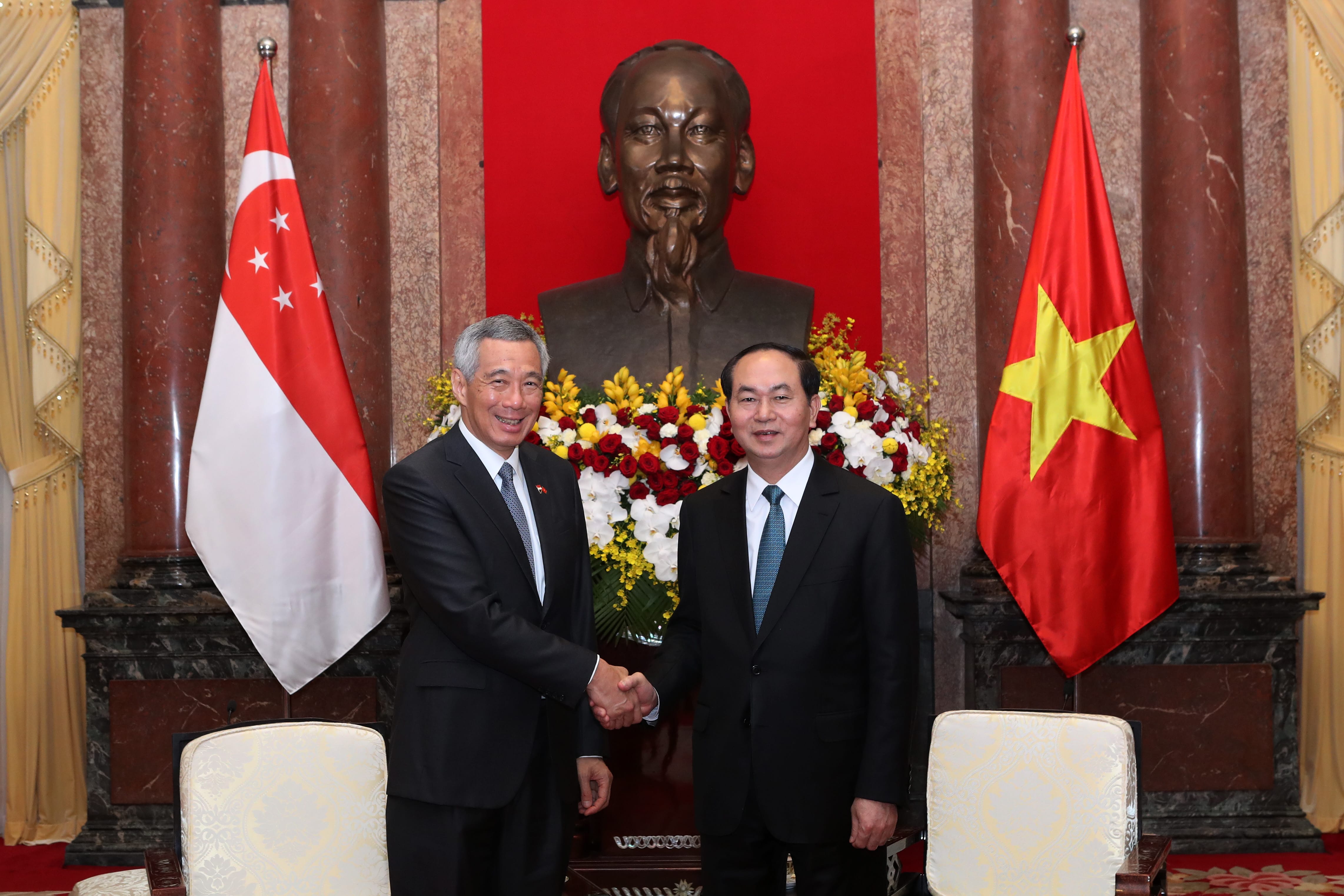 Two men in suits shake hands between Singapore and Vietnam flags, under a bronze bust.