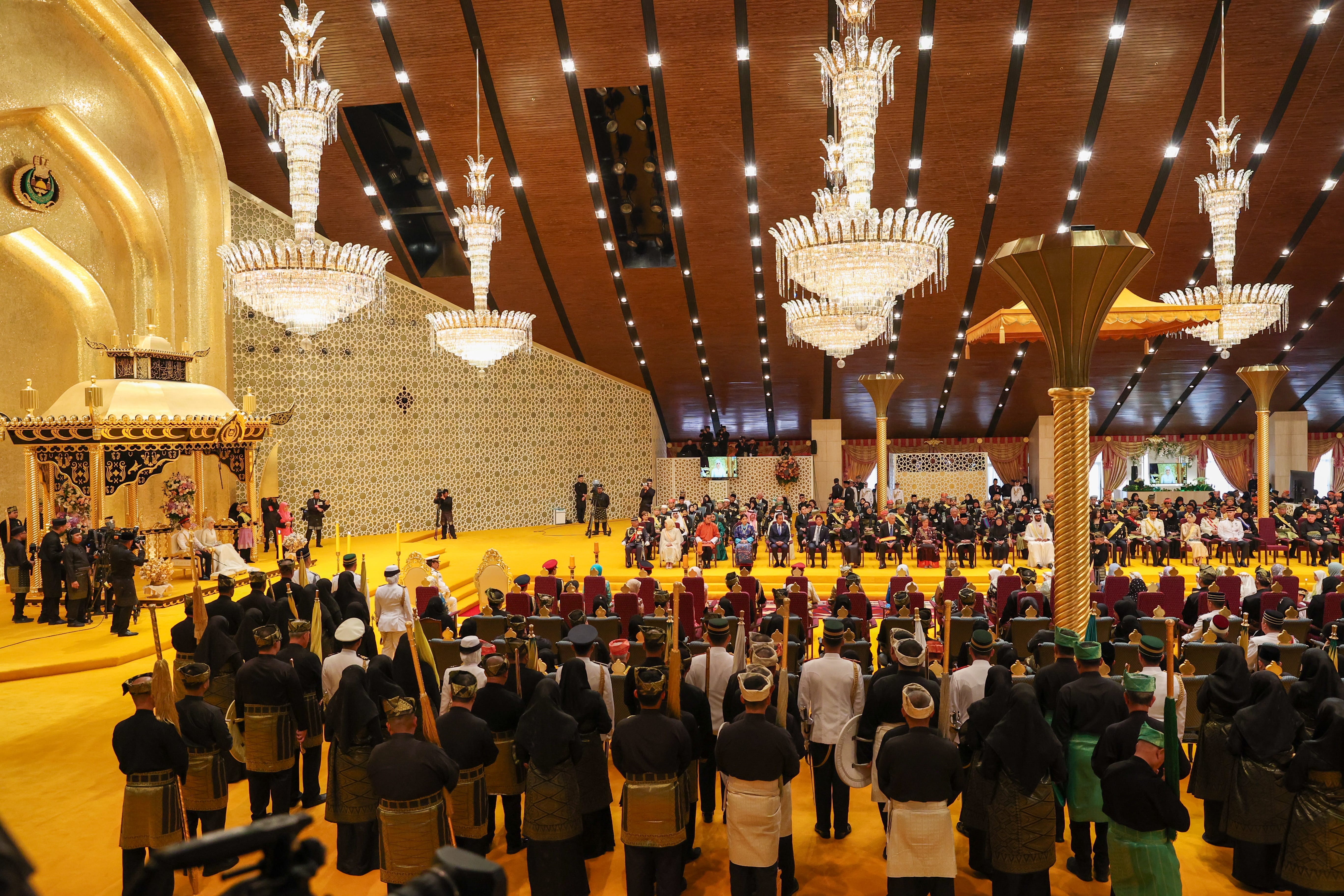Large formal hall with seated crowd facing a Sultan on a golden throne.