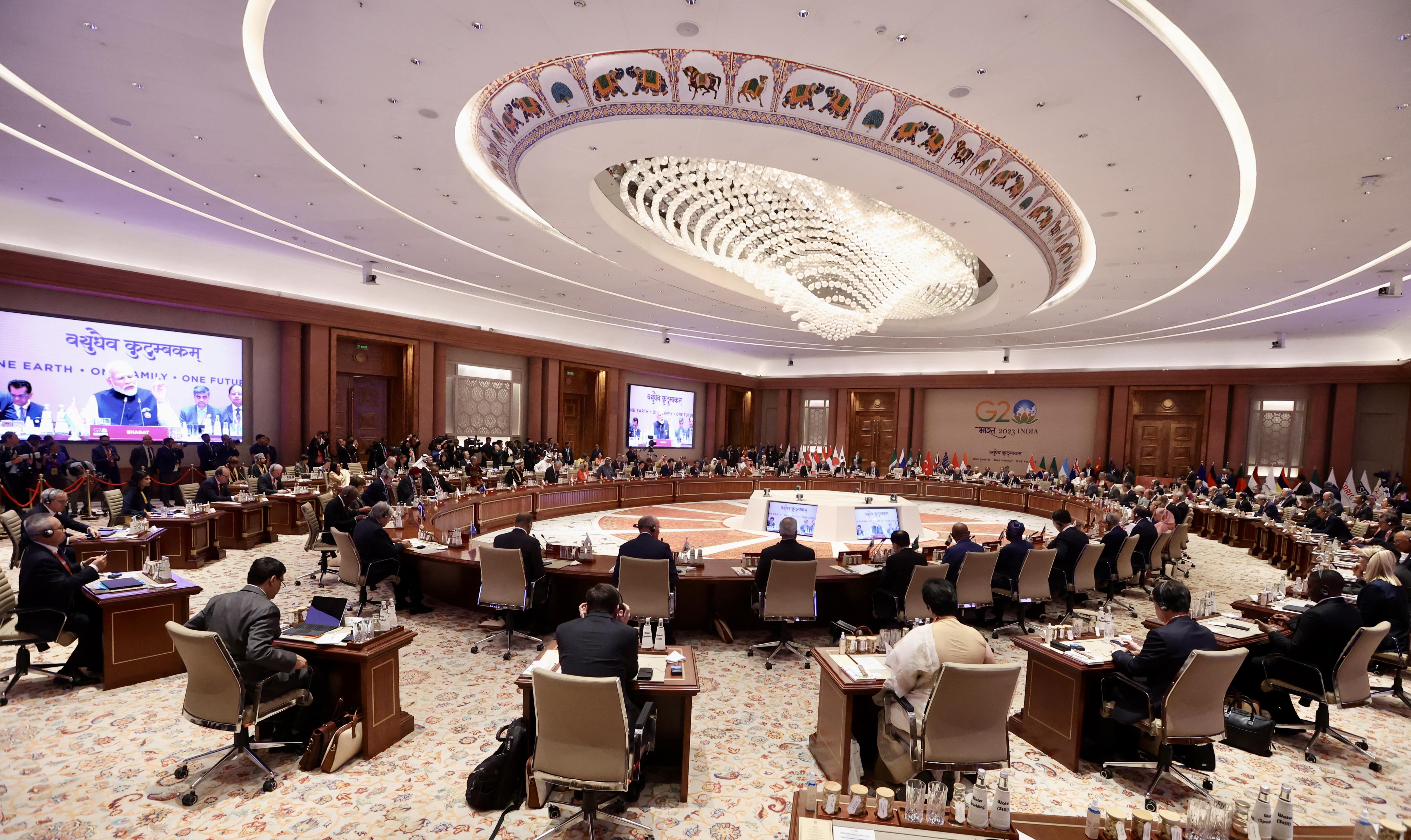 People at G20 India meeting, seated around a large round table, flags displayed.