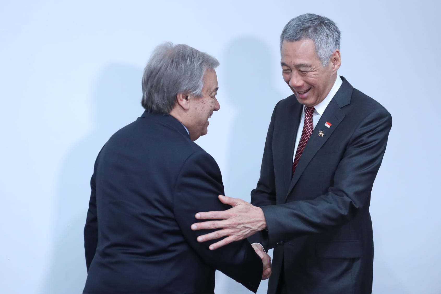 Antonio Guterres and Lee Hsien Loong in suits, shaking hands against a white wall.
