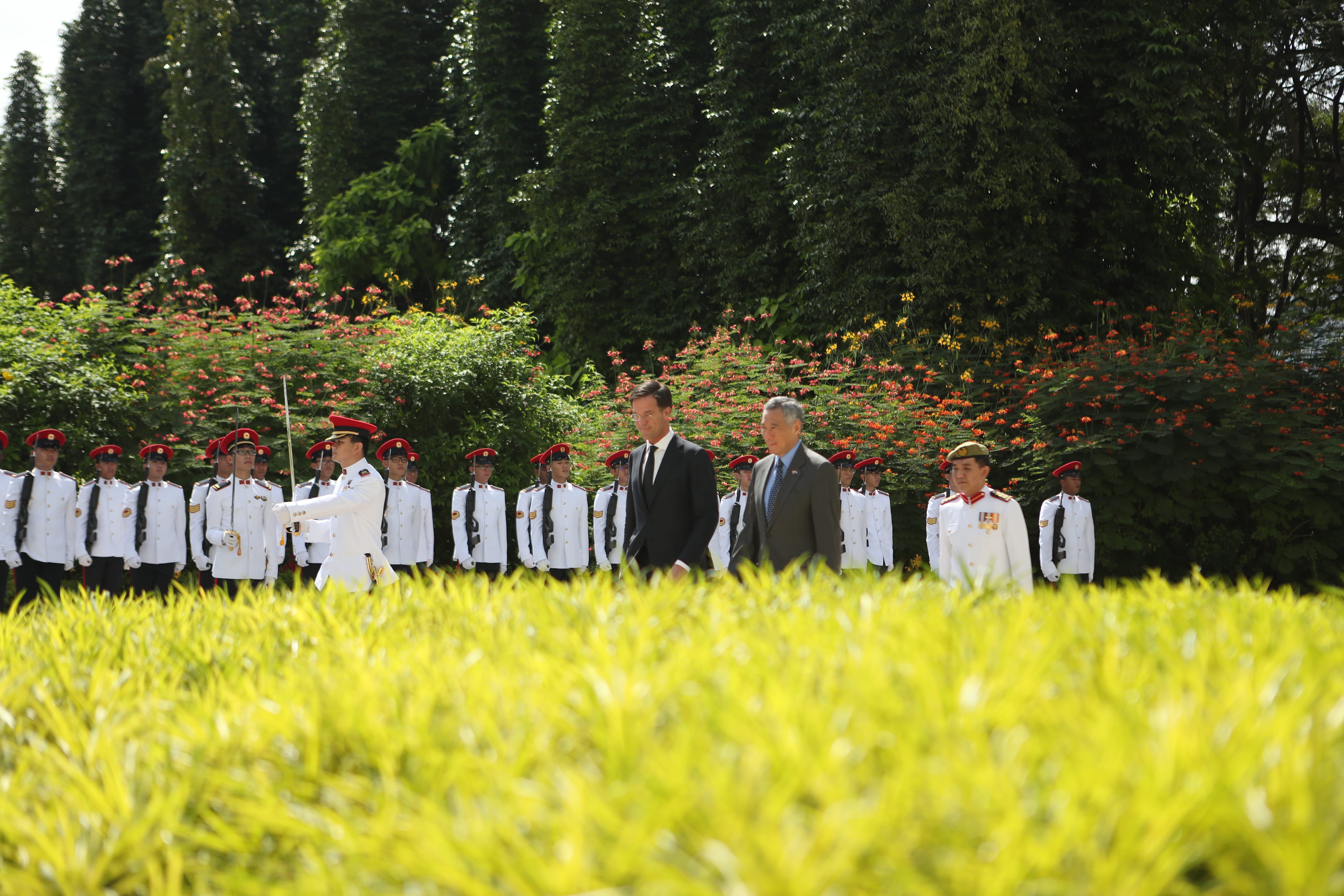 Two men in suits walk past guards in white uniforms with rifles and red hats.
