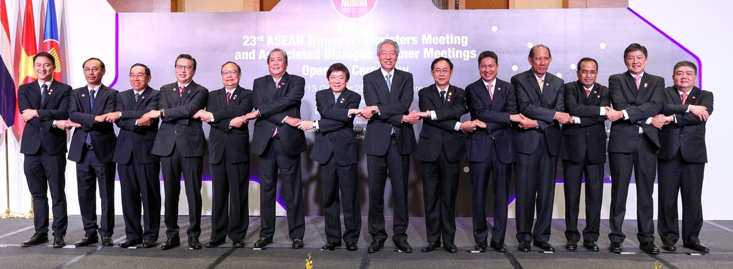 Thirteen people in suits link arms on a stage in front of the ASEAN Transport Ministers Meeting banner.