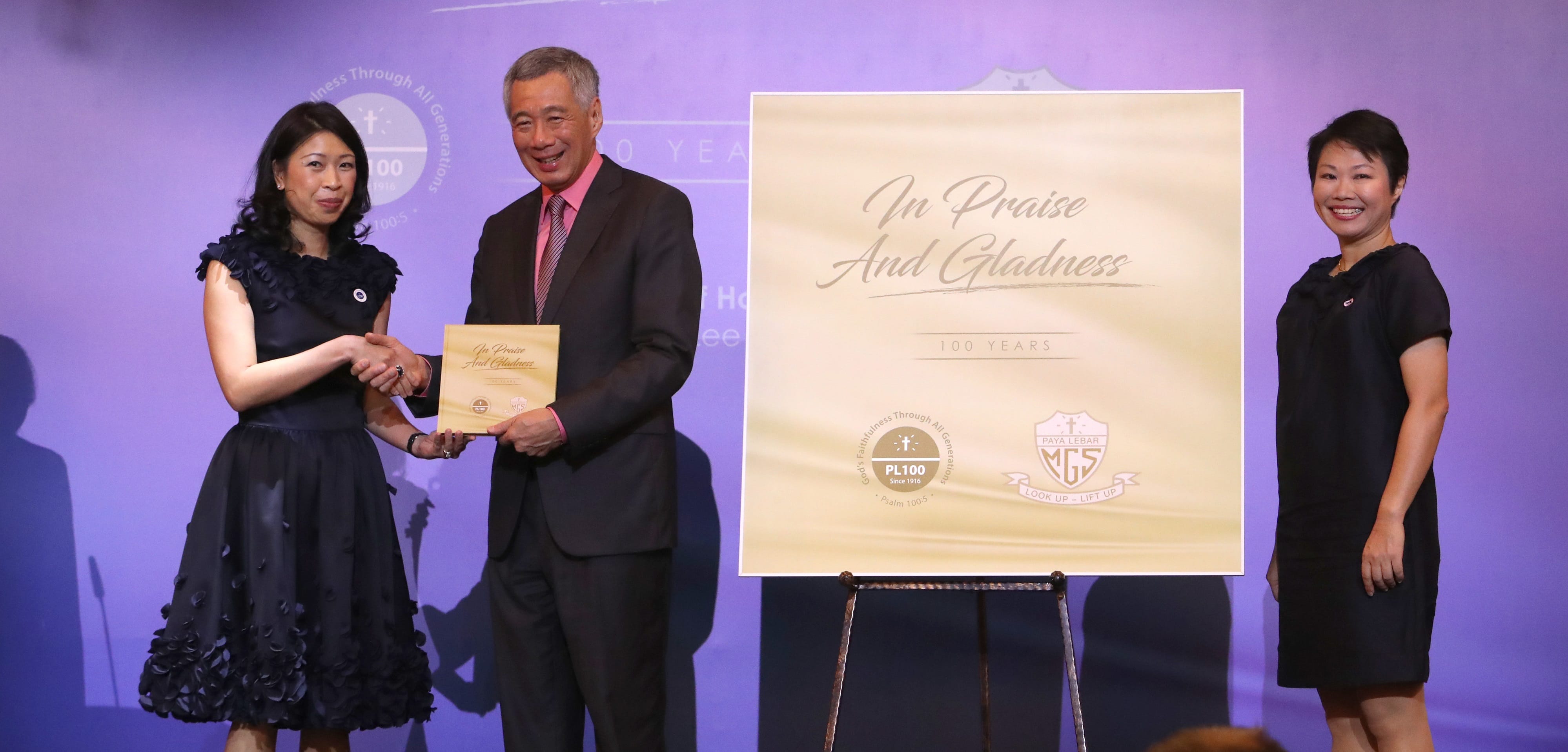 Two women and Singapore's Prime Minister, Lee Hsien Loong, onstage with "In Praise and Gladness" poster.