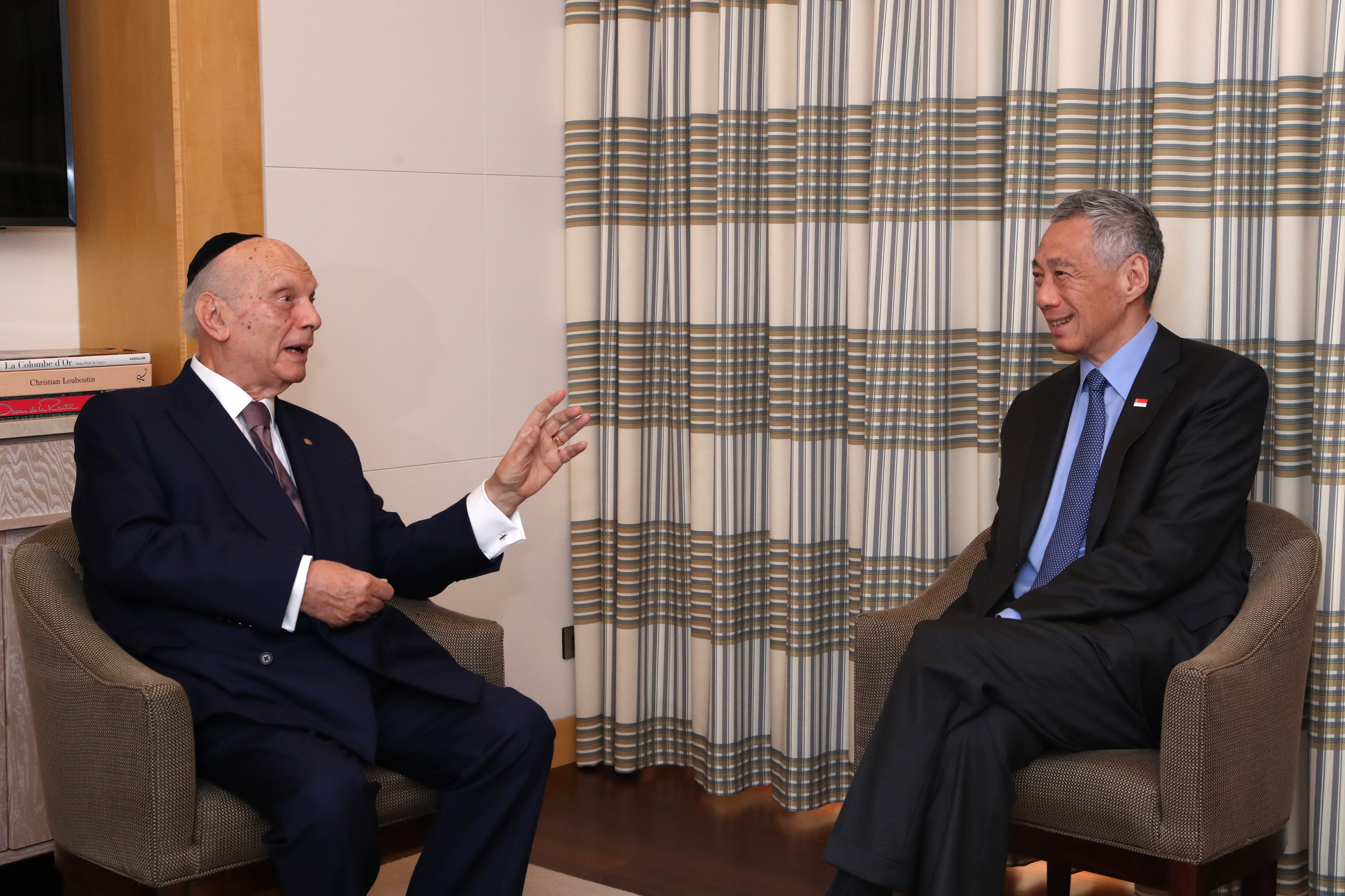 Two men in suits sit in chairs. One gestures with his hand. The other has a small Singapore flag pin.