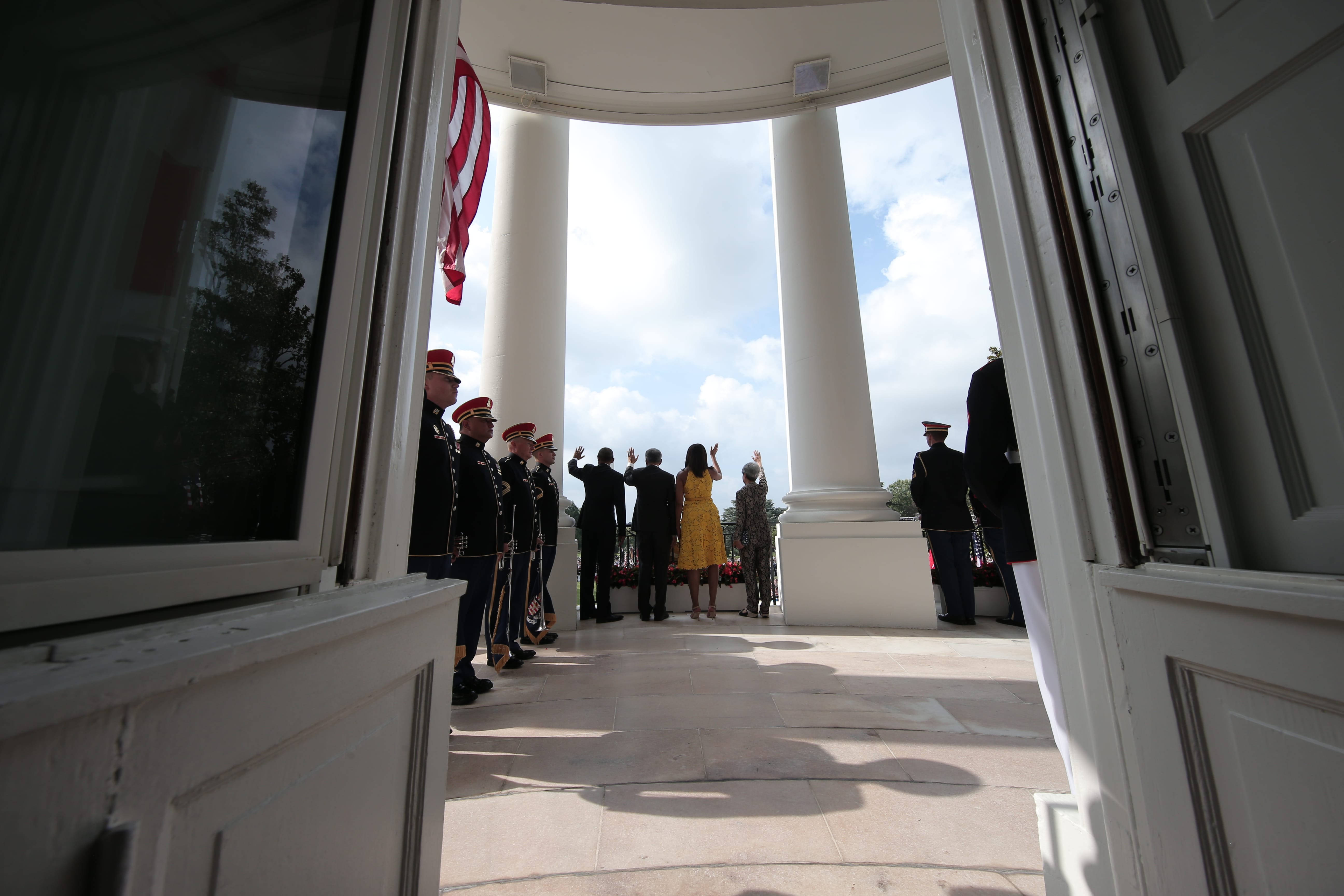 White House portico framed by doorway, people waving, U.S. flag, guards in uniforms.