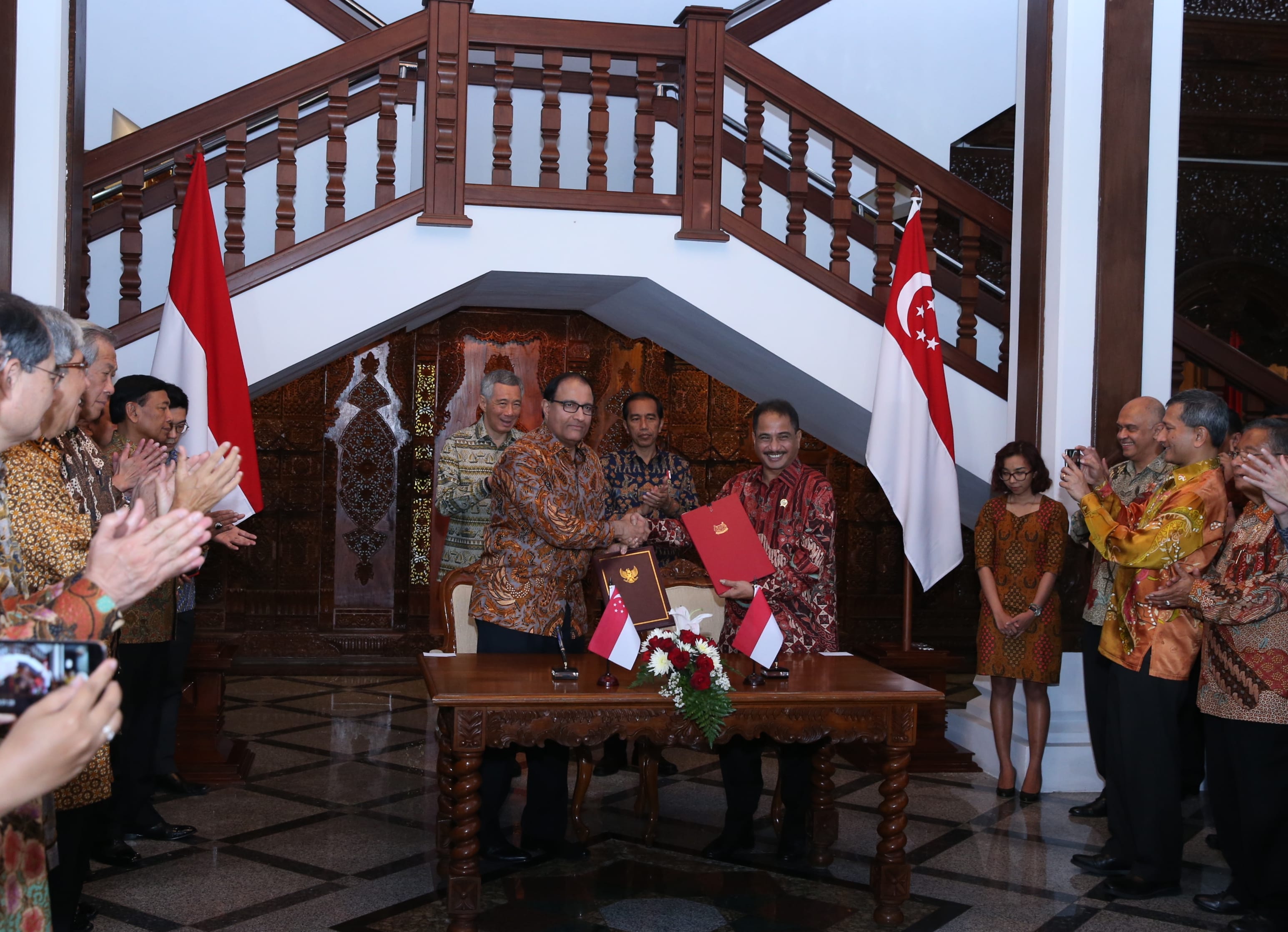Two men shake hands across a table with flags of Indonesia and Singapore present. People applaud in the background.