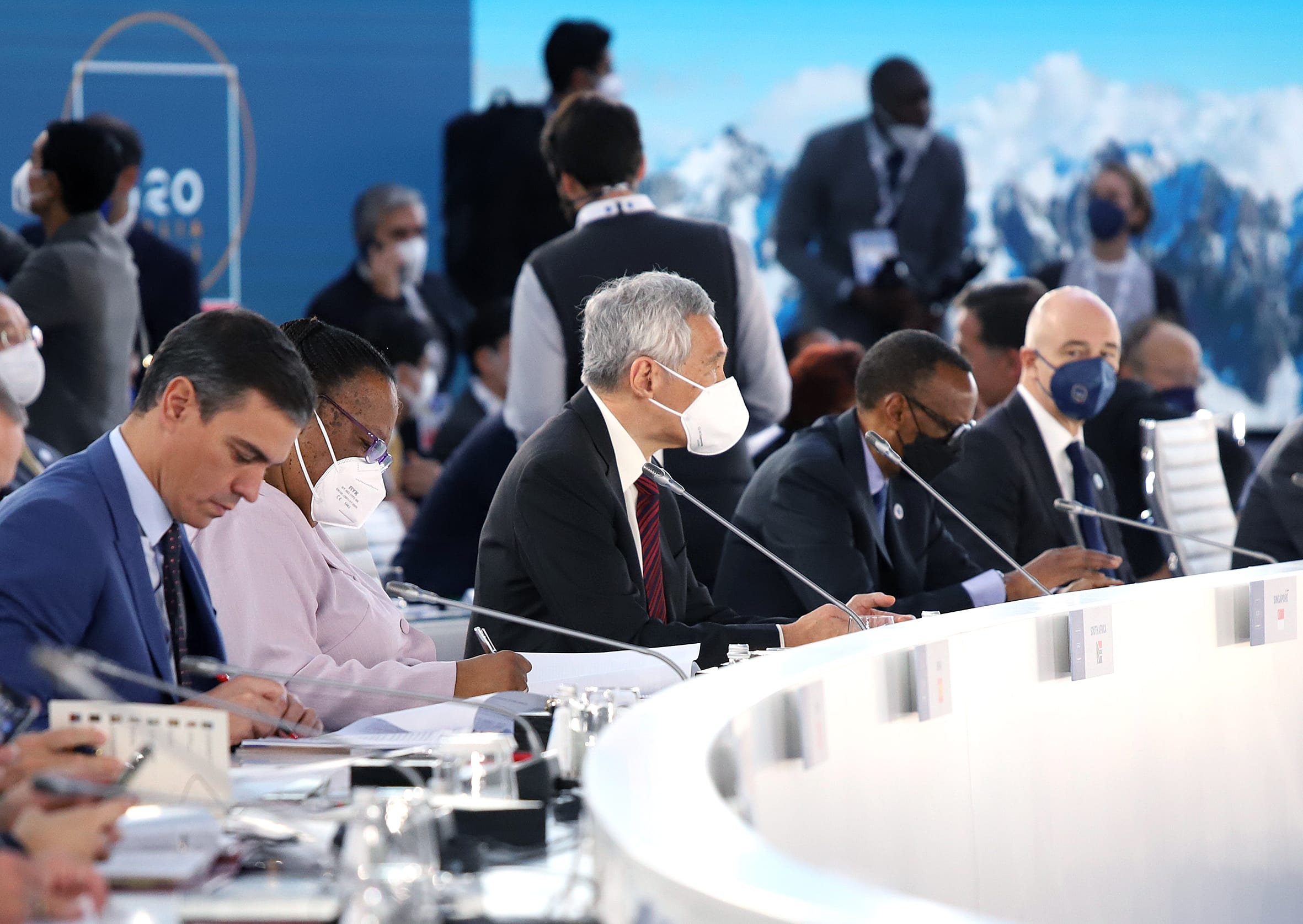 Attendees wearing masks at G20 summit, some writing, with country name cards on the table.