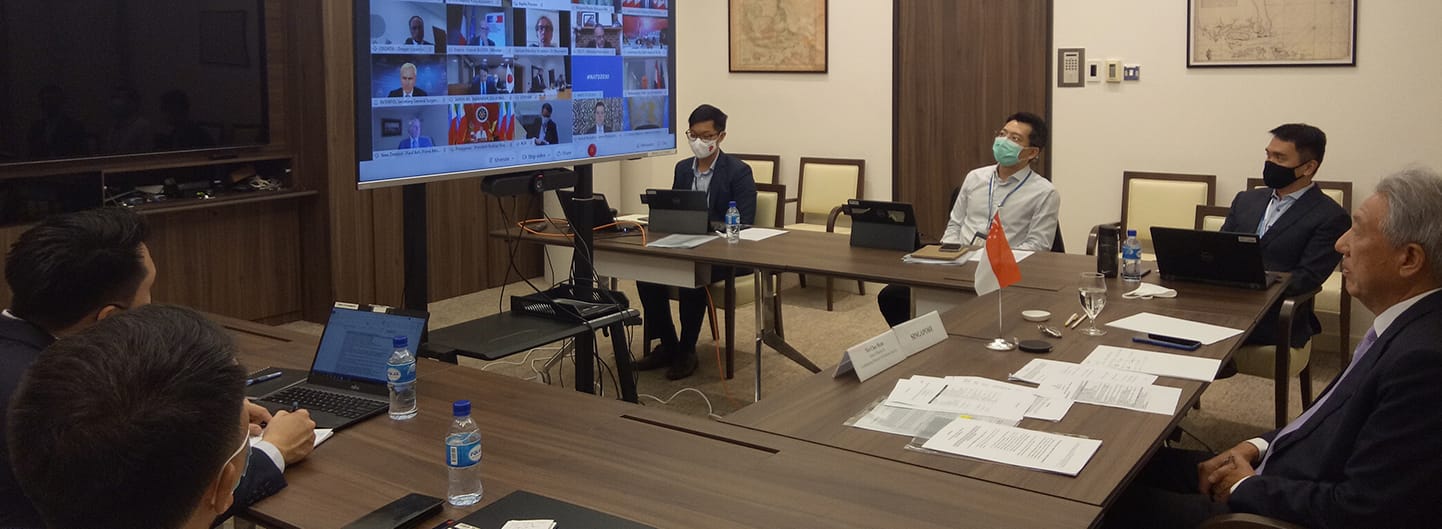 People wearing masks at a U-shaped table with laptops, watching a video conference.