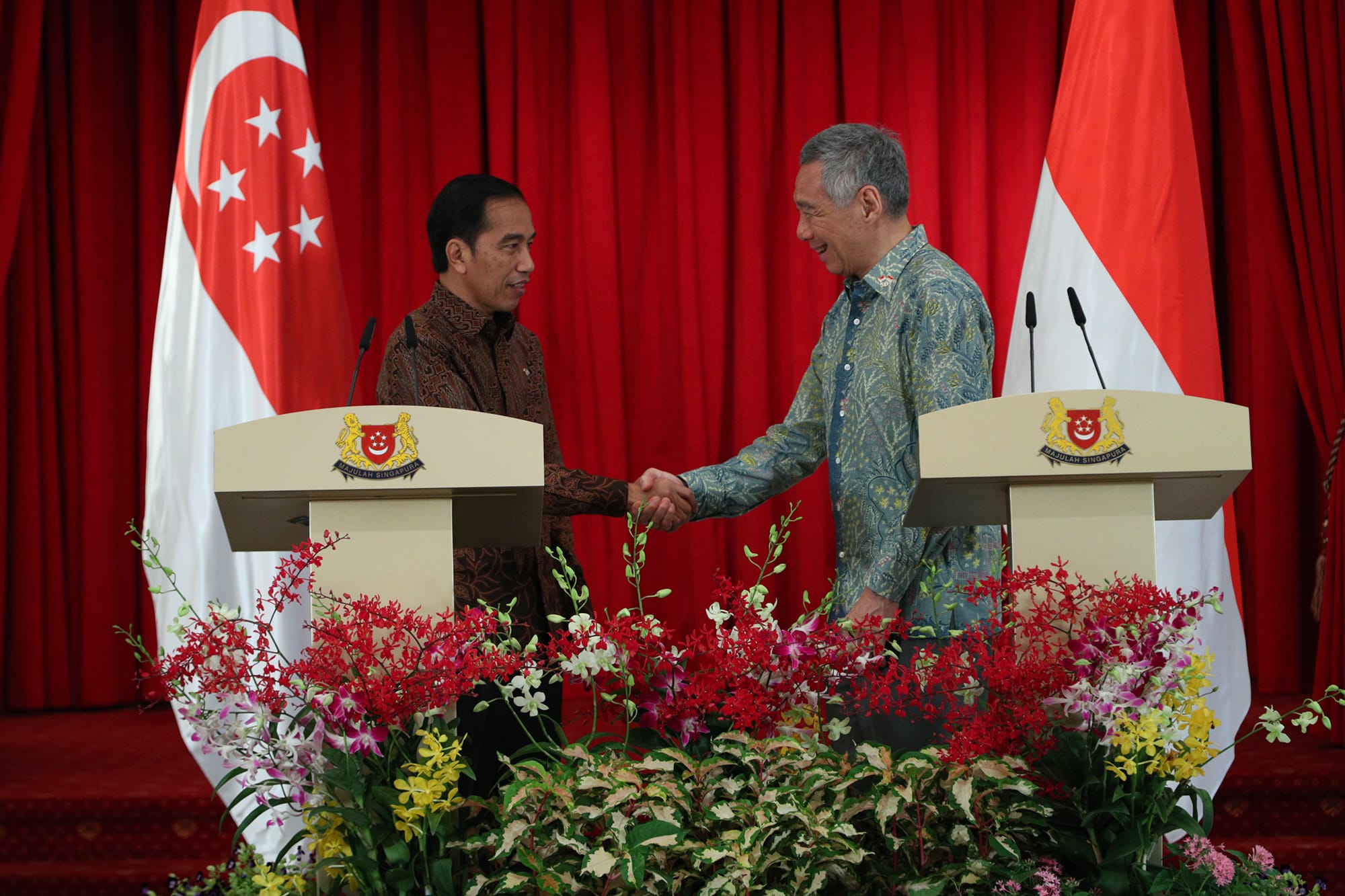 Two men shake hands at lecterns with Singapore flags, behind red curtains and floral arrangements.
