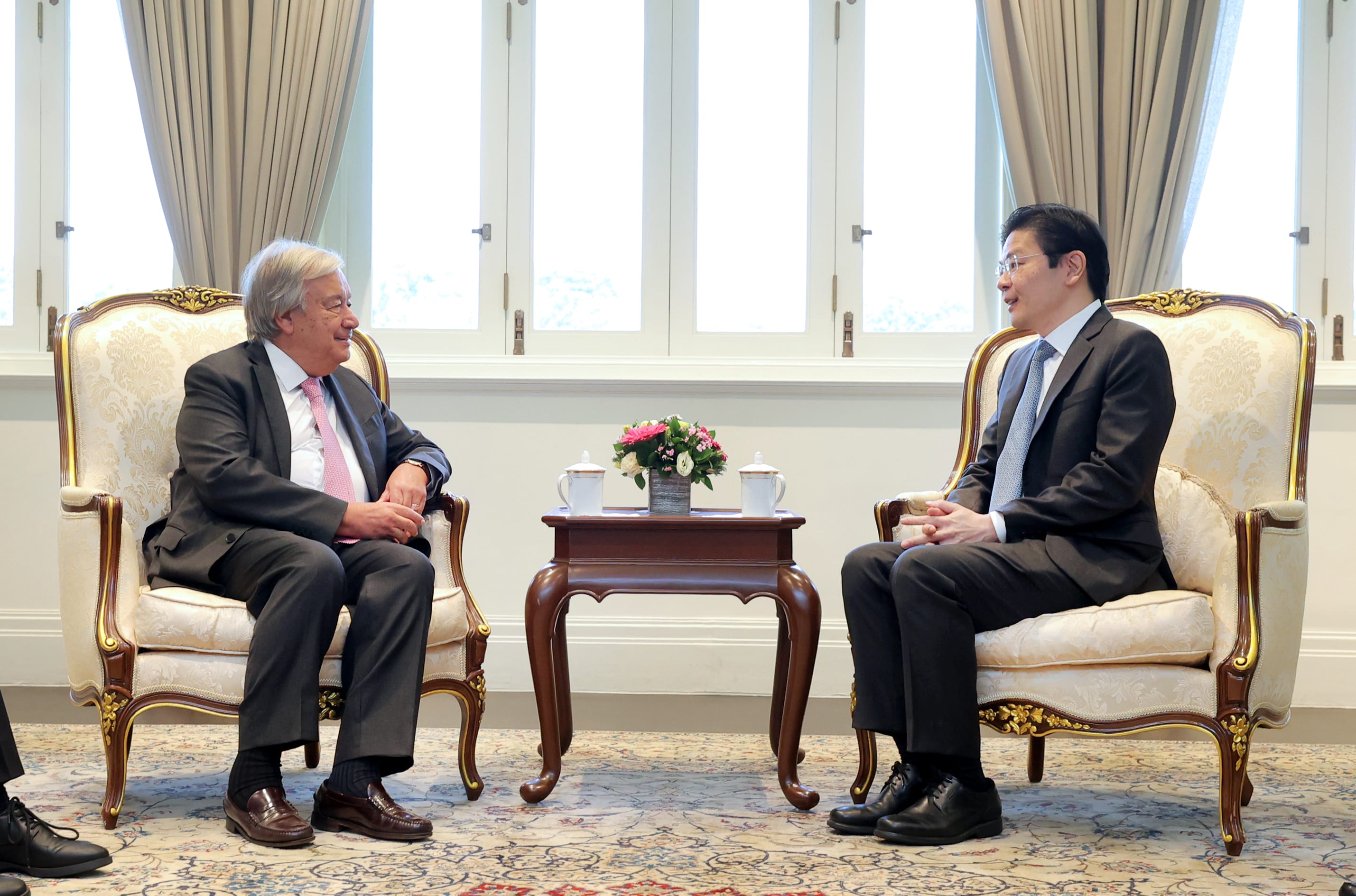 António Guterres and a man in suits sit in ornate chairs facing each other in front of a window.