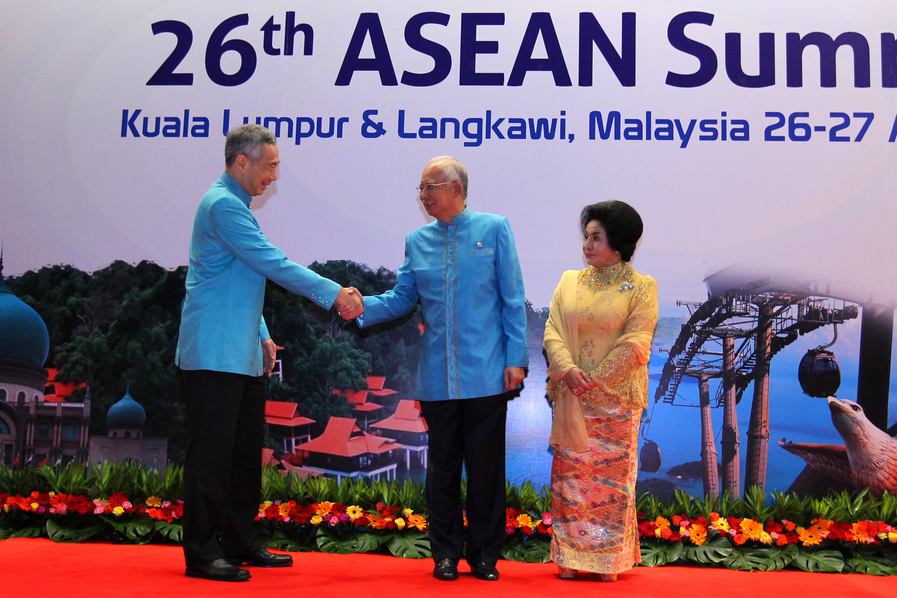Two men in blue shake hands; woman in gold stands next to banner for 26th ASEAN Summit.