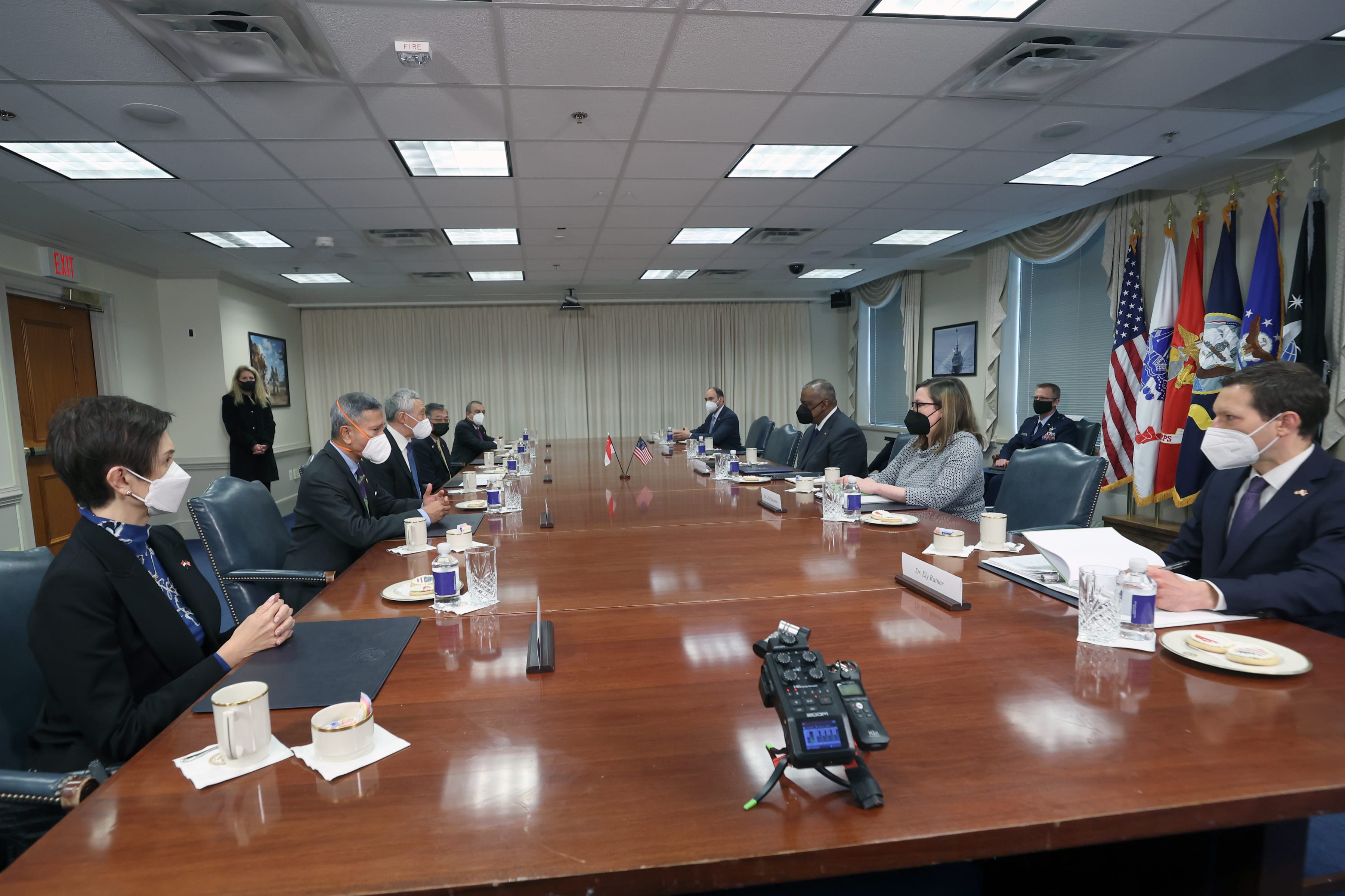 People with face masks seated around a long conference table. Flags are visible in the background.