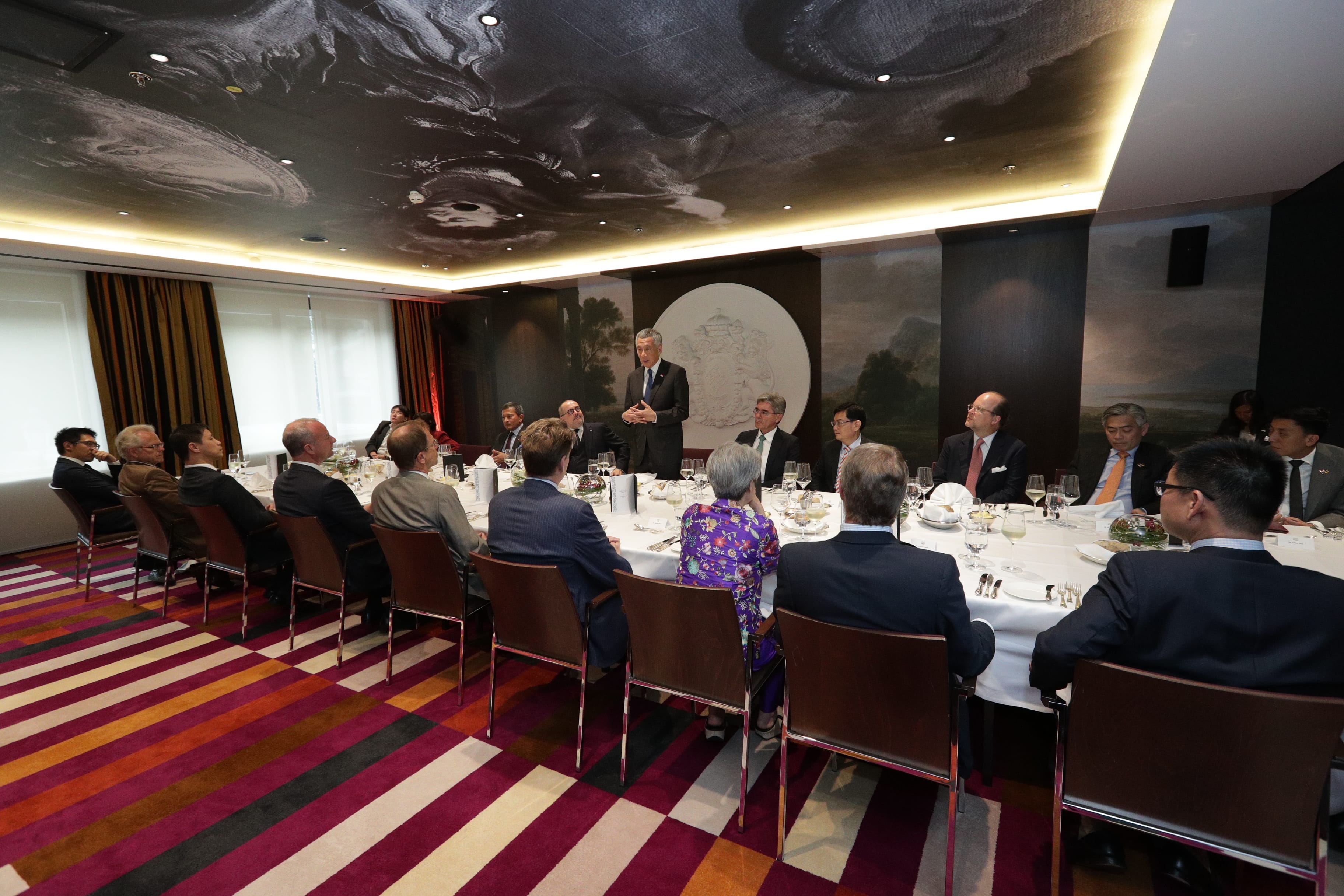 Lee Hsien Loong speaks at a long table with attendees in suits. Wall art, ceiling with cloud design.