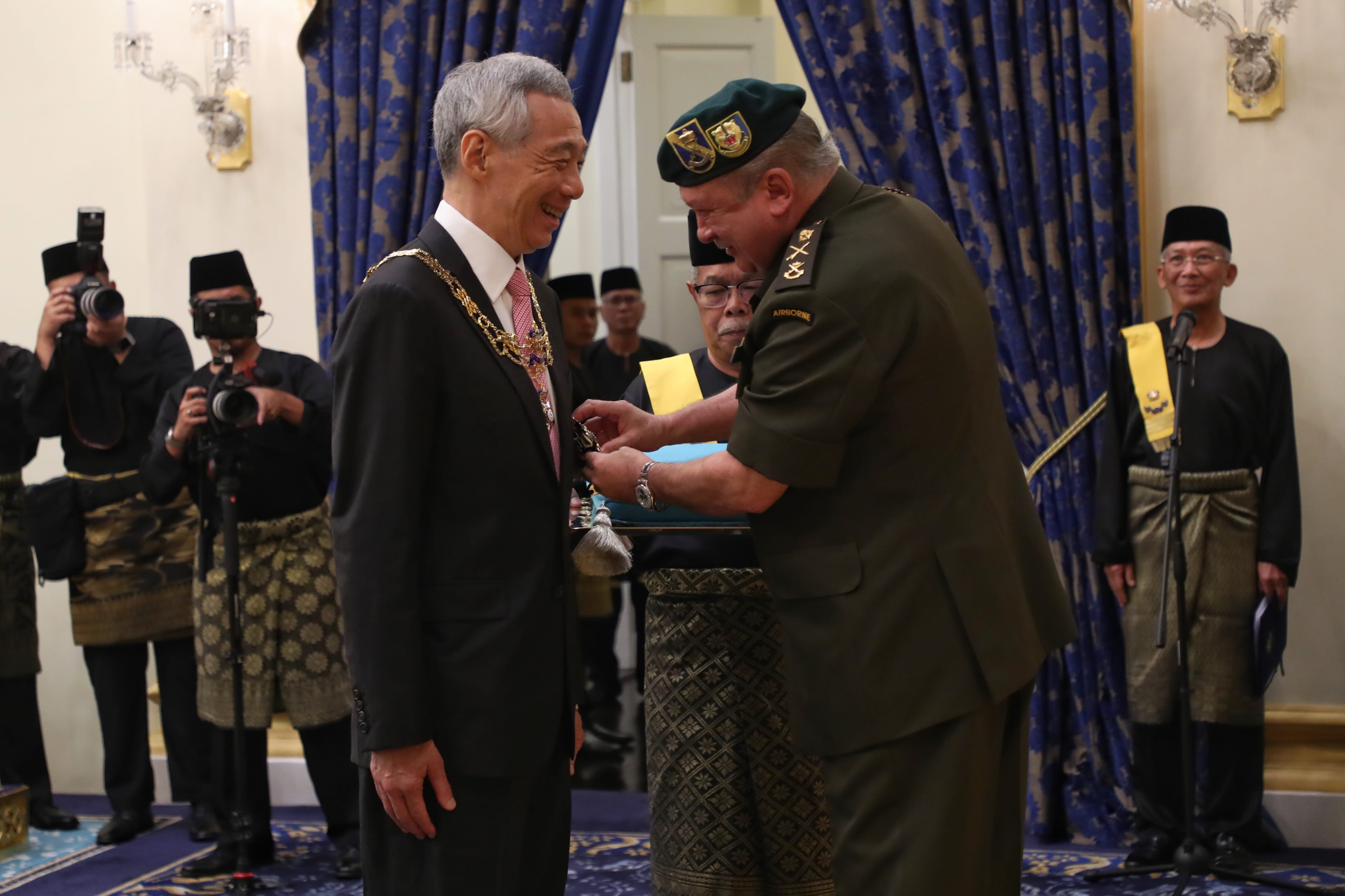 Man in military uniform pinning medal on Lee Hsien Loong, surrounded by photographers and officials.