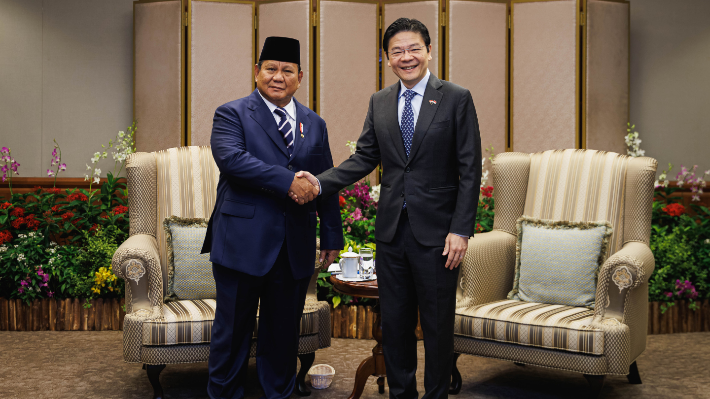 Two men in suits shake hands in front of ornate chairs and floral arrangements.