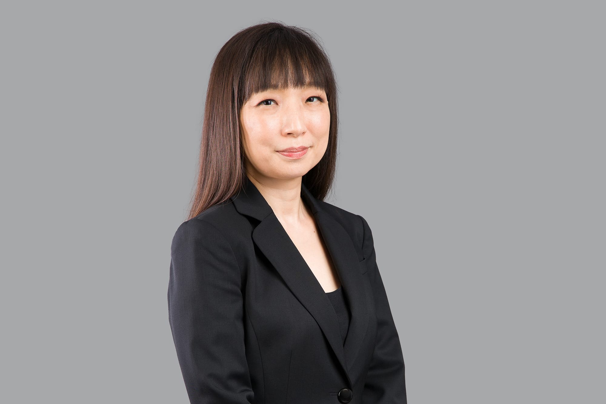 Asian woman in black suit, long brown hair with bangs, against a gray background.