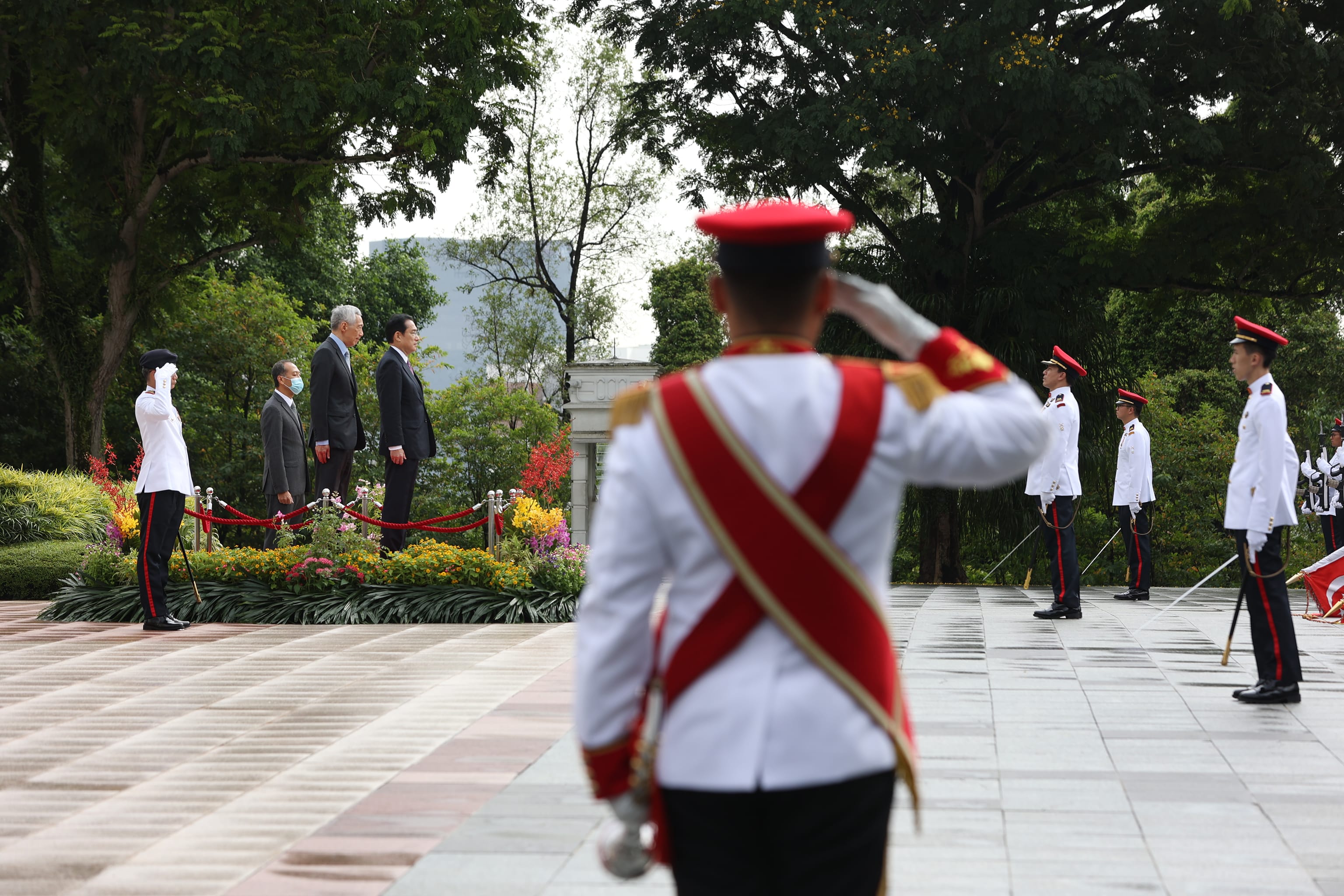 Honor guard in white uniforms, red hats, and sashes salute three men in suits. Outdoors, park background.